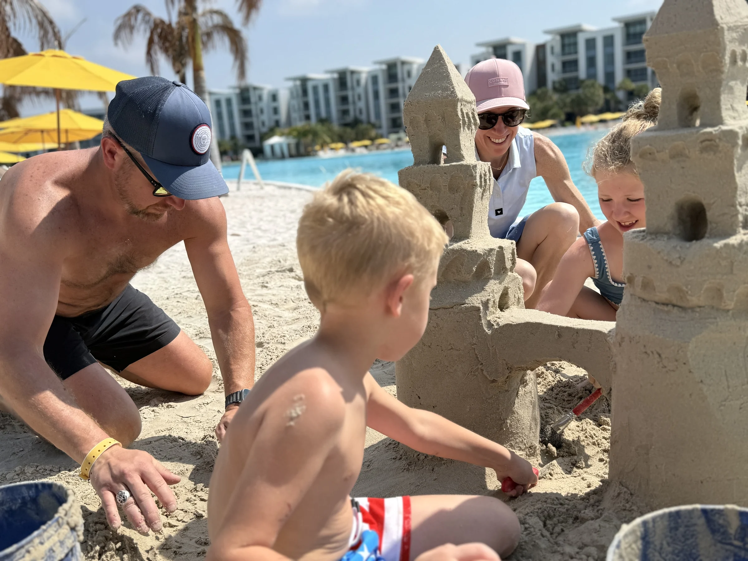 Family building detailed sandcastles on a sandy beach near a pool with yellow umbrellas and buildings.