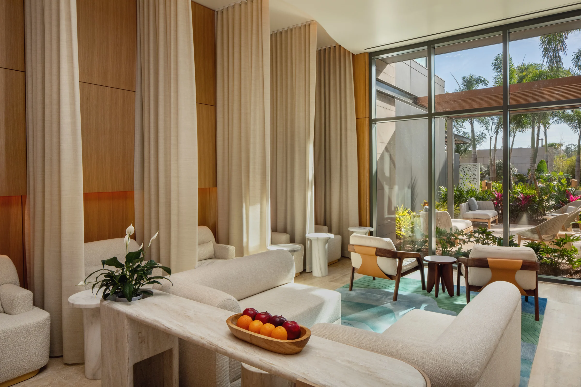 Modern lounge with beige seating, wooden accents, marble tables, and large glass windows overlooking a garden.