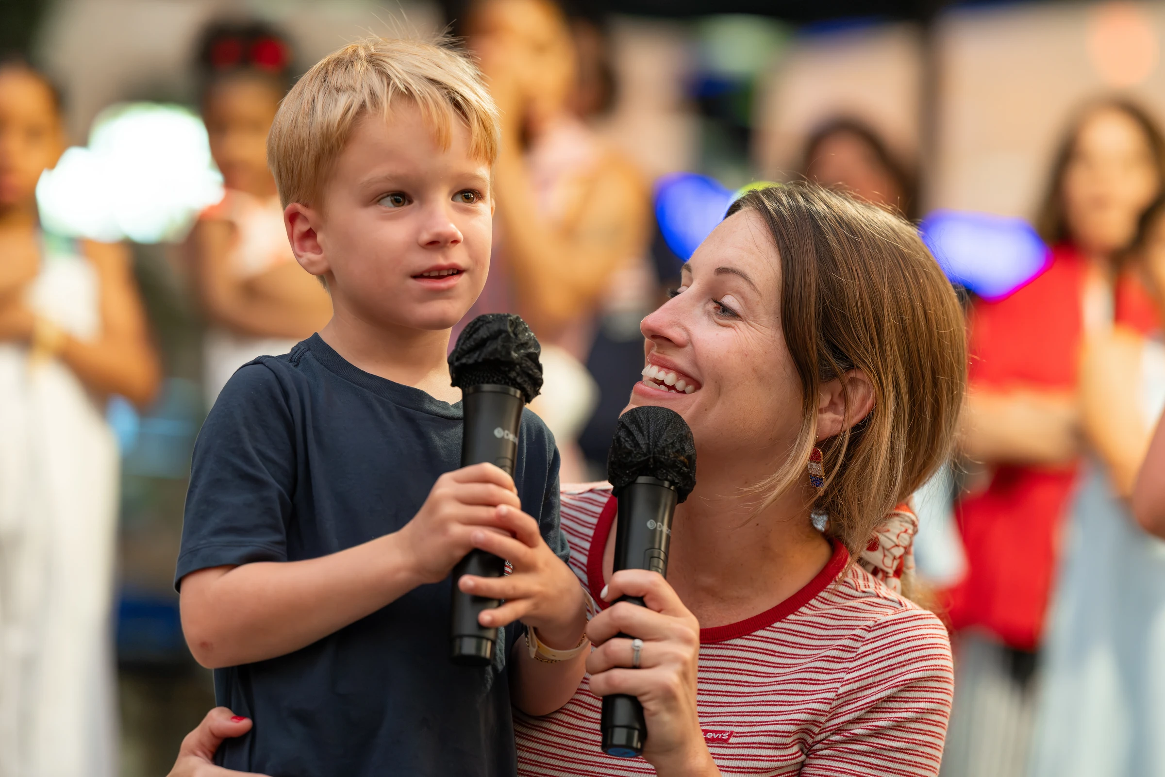 Smiling woman holding a microphone looks at young boy holding another microphone at a public event.