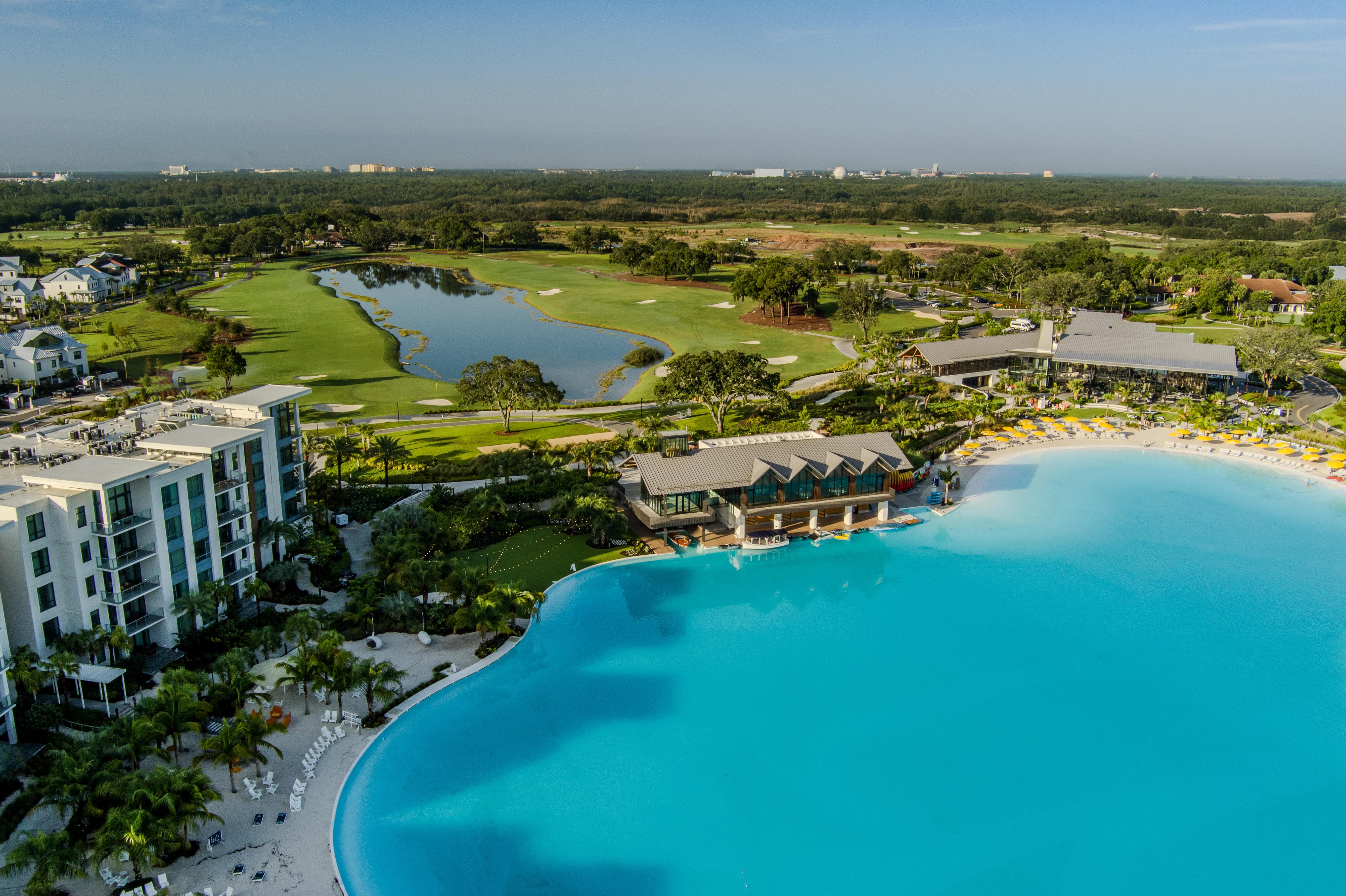 Large resort pool with yellow umbrellas, nearby modern buildings, golf course, and forest in the distance.