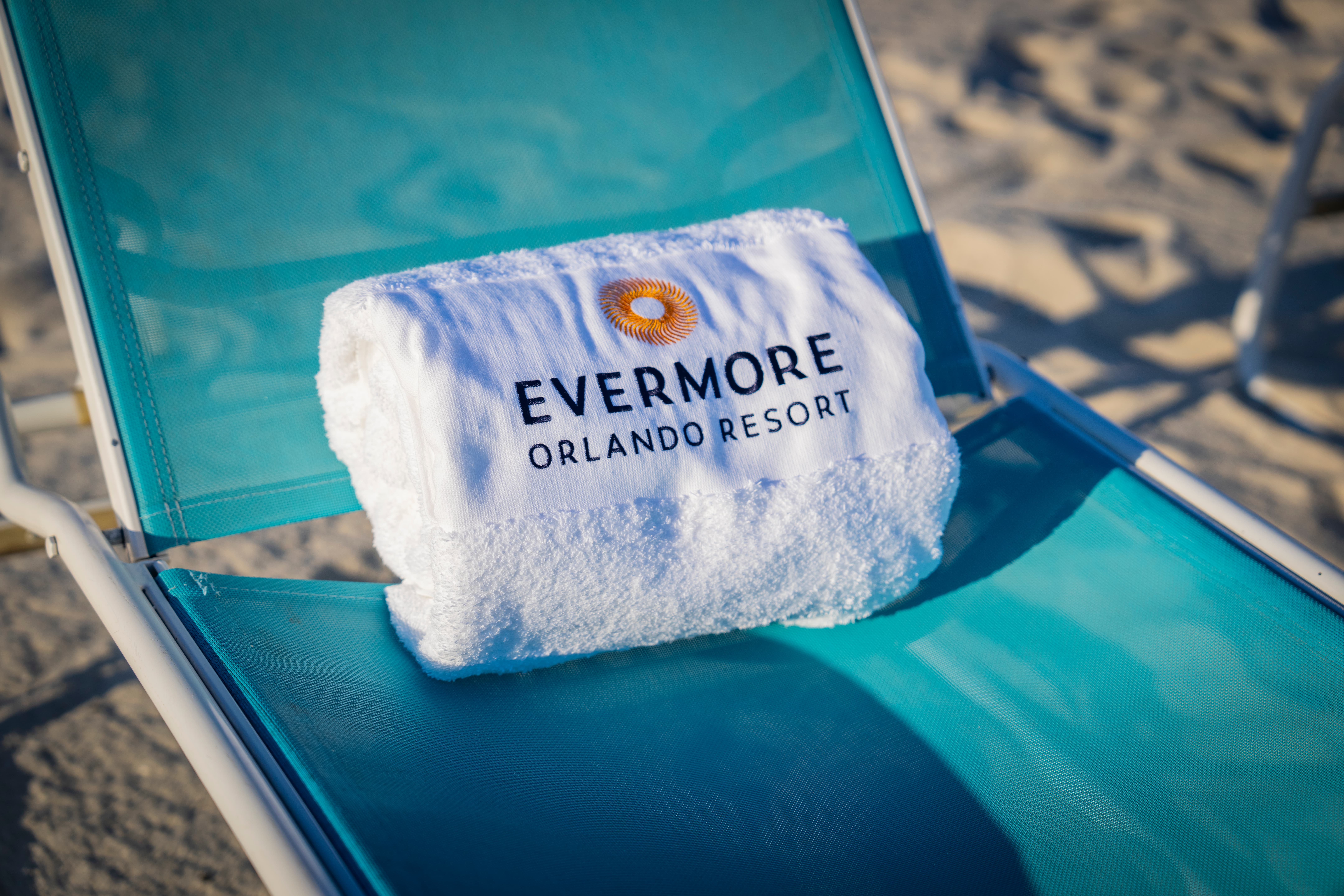 White folded Evermore Orlando Resort towel on a teal beach chair with sand in the background.