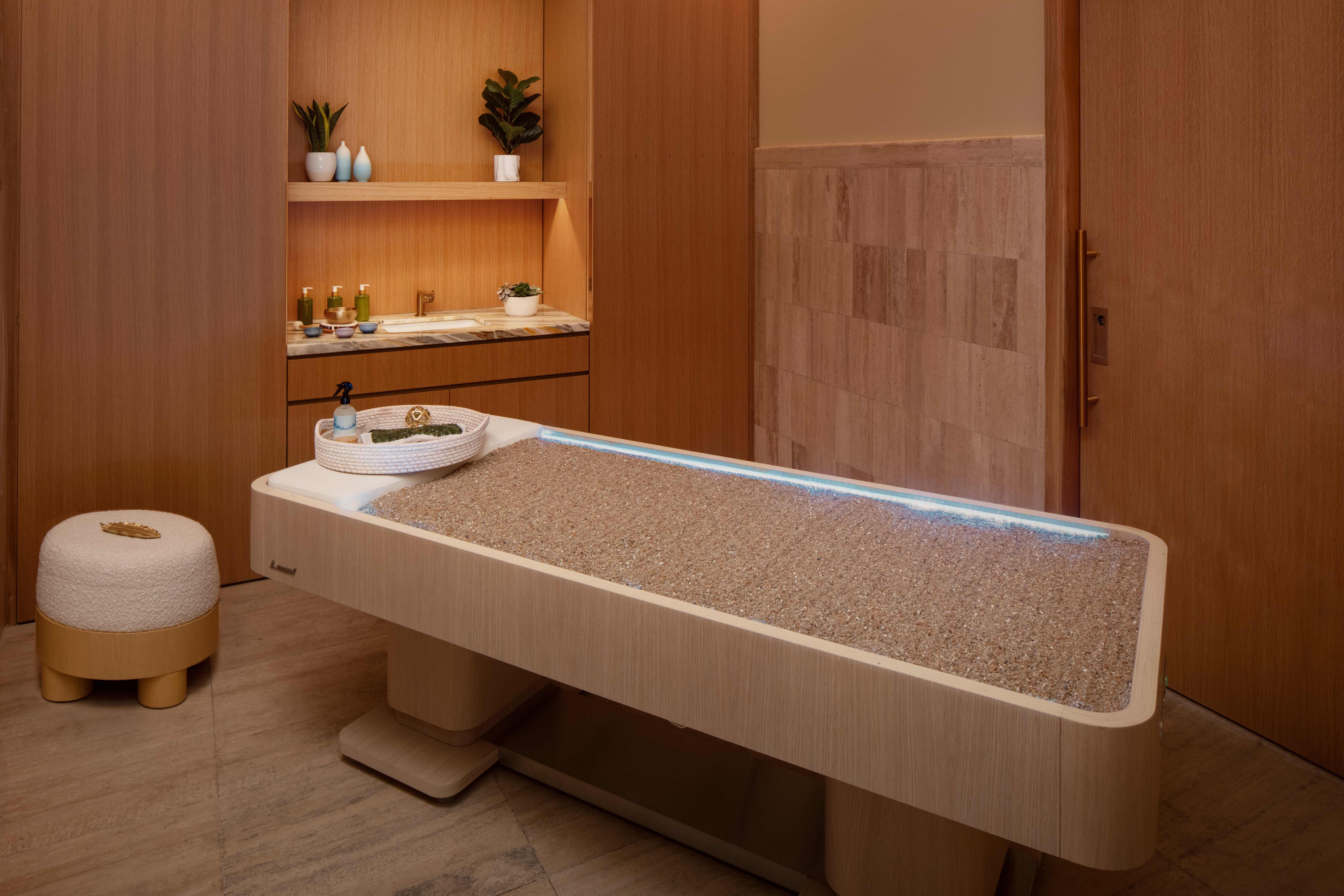 Minimalist spa treatment room with wooden décor, pebble massage table, and plants on shelves.