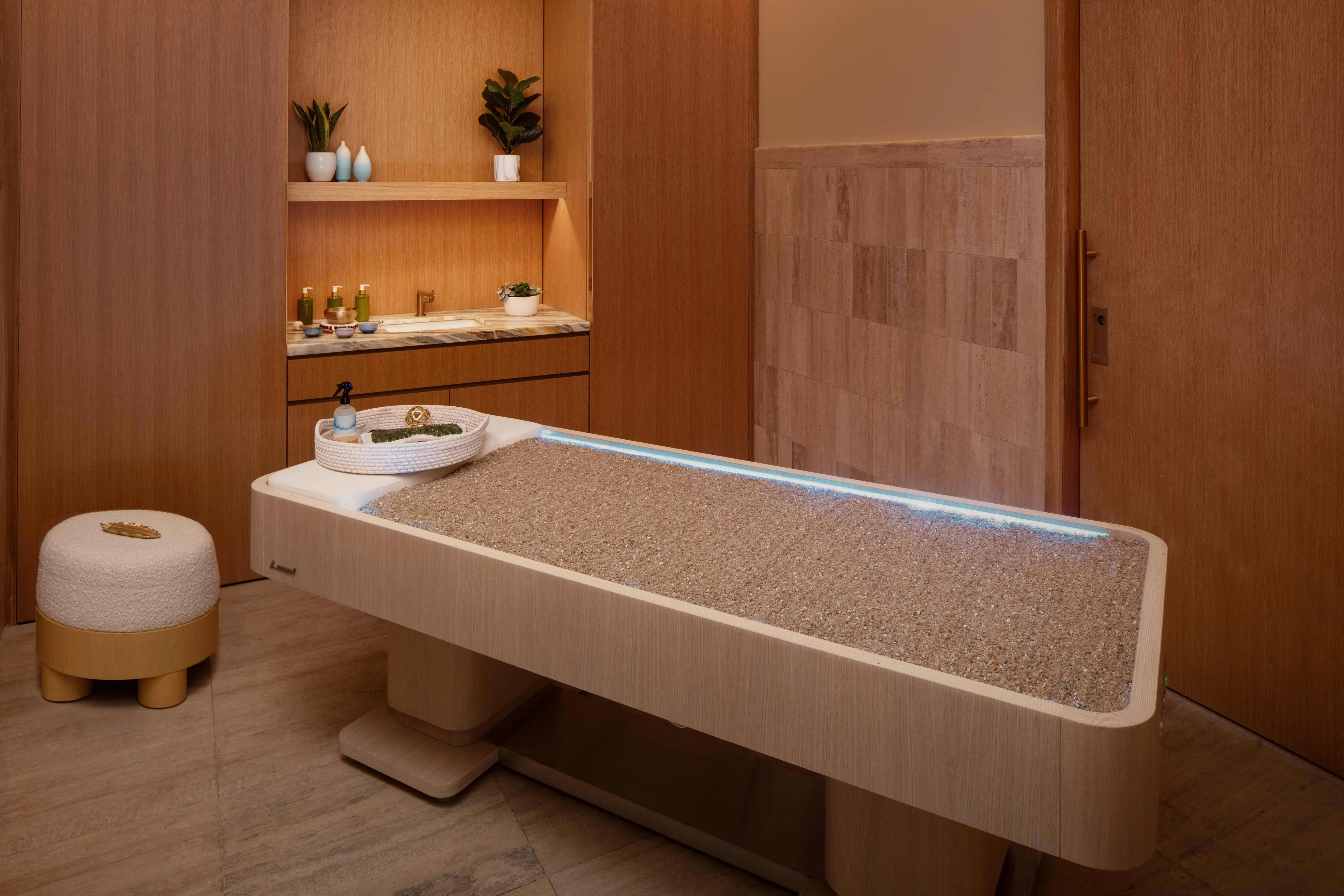 Minimalist spa treatment room with wooden décor, pebble massage table, and plants on shelves.