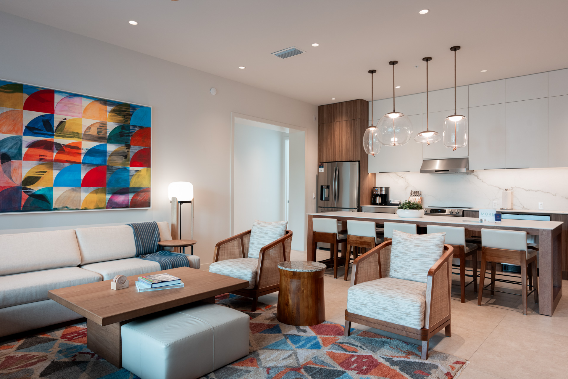 Modern open-concept living room and kitchen with beige sofa, armchairs, wooden table, and colorful rug.