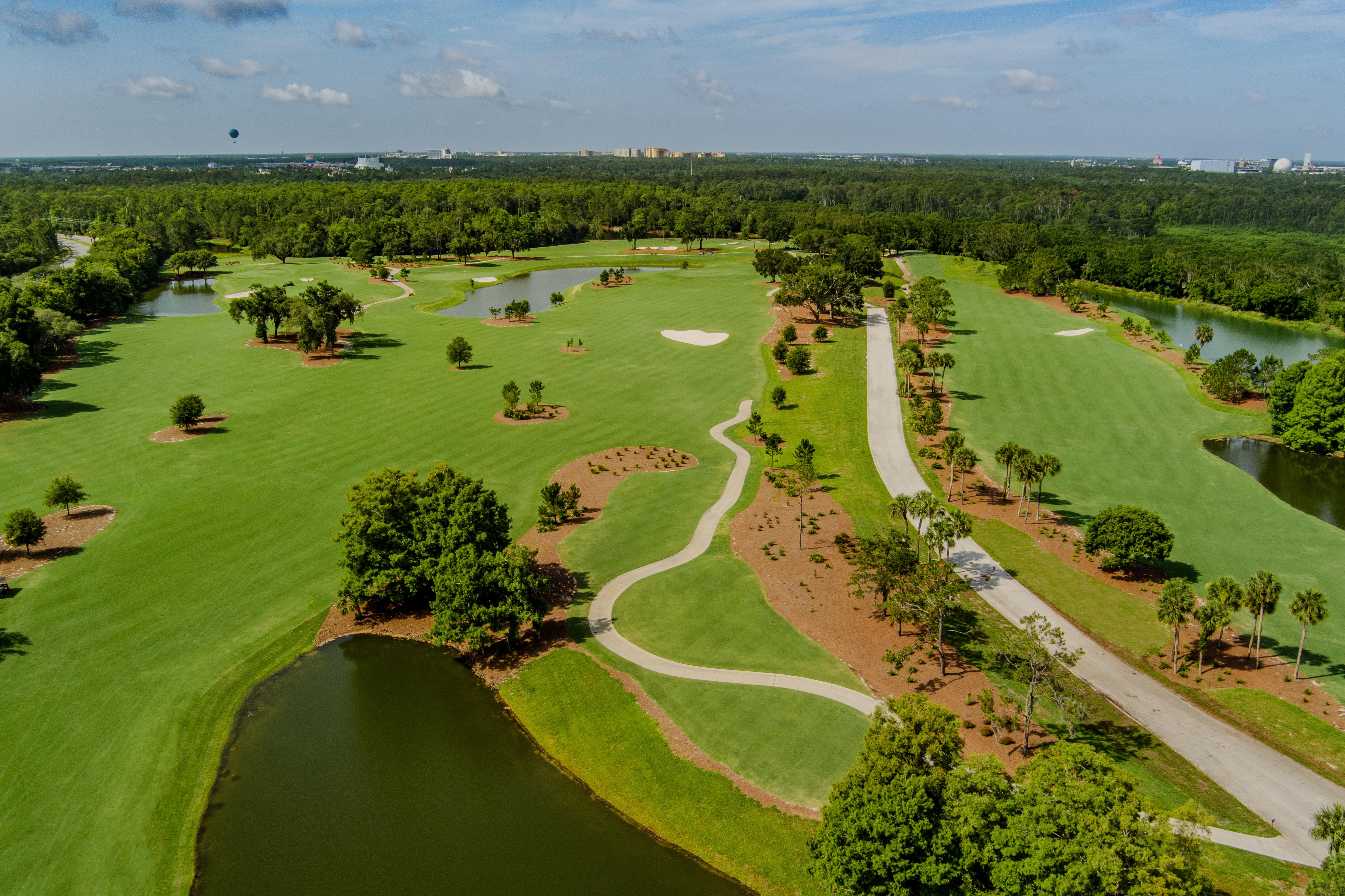 Aerial view of a green golf course with trees, ponds, paths, and sand bunkers under a blue sky.