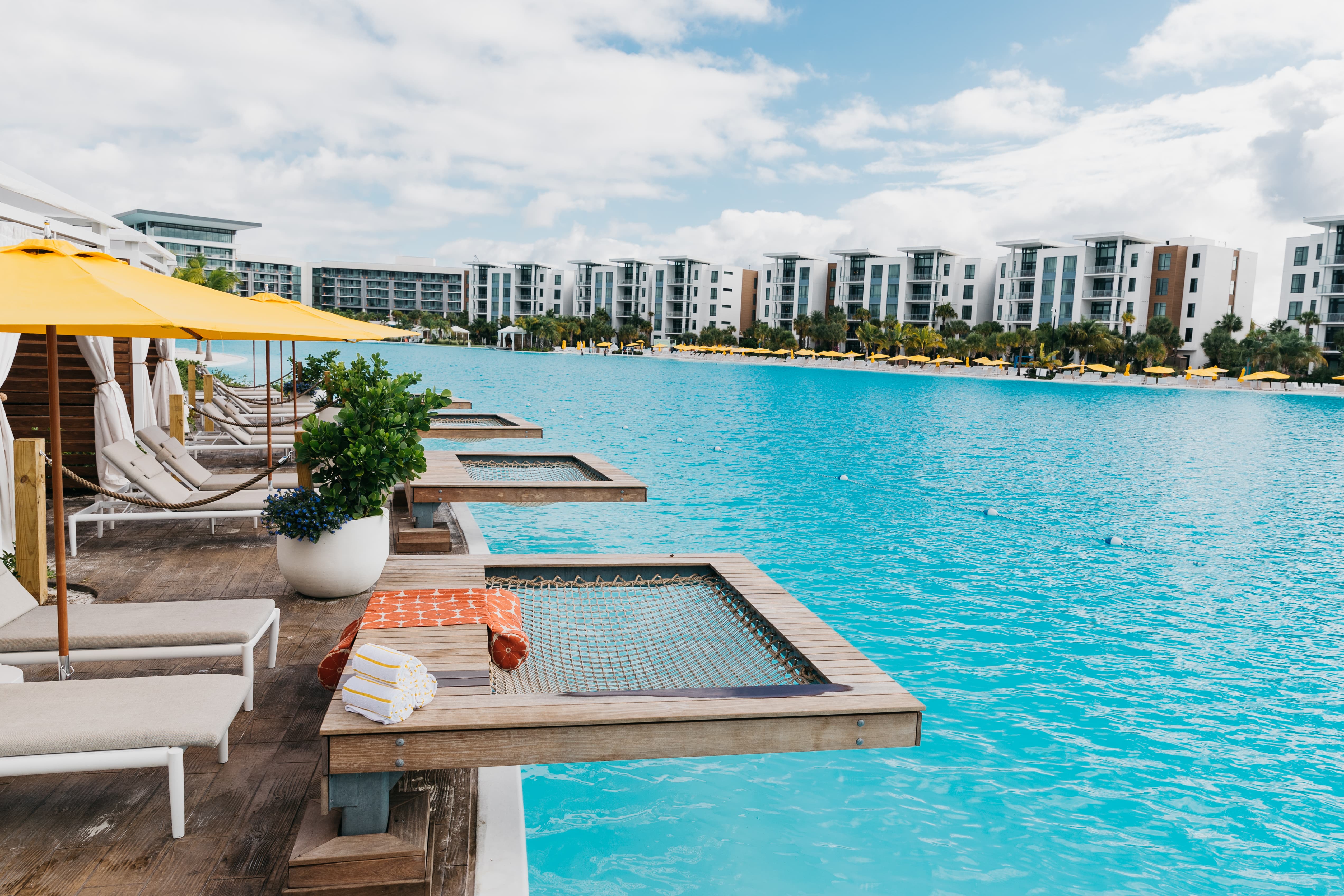 Luxury poolside with wooden net loungers, yellow umbrellas, and modern white buildings in the background.