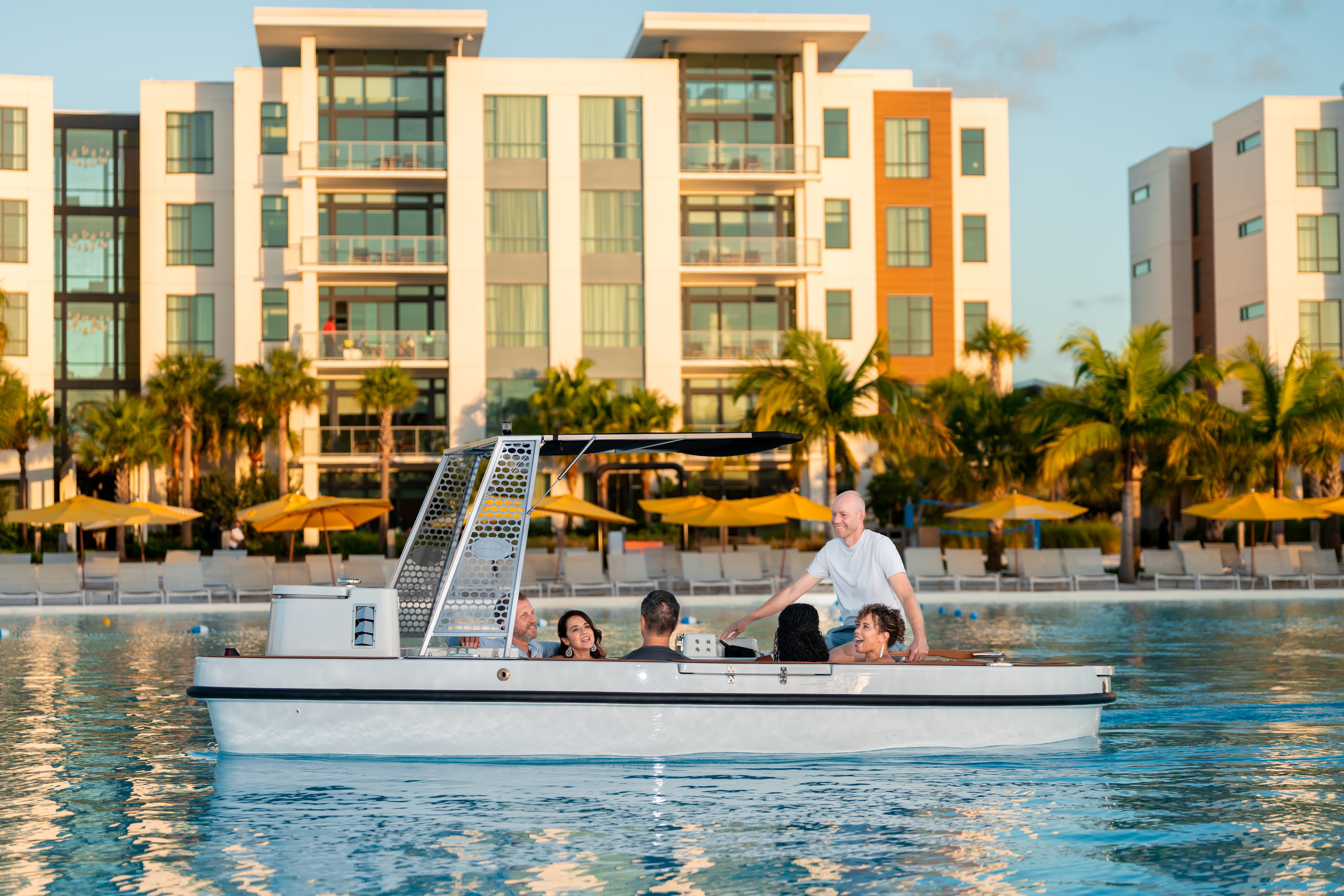 Group of six adults riding a white electric boat on a pool with buildings and palm trees behind.