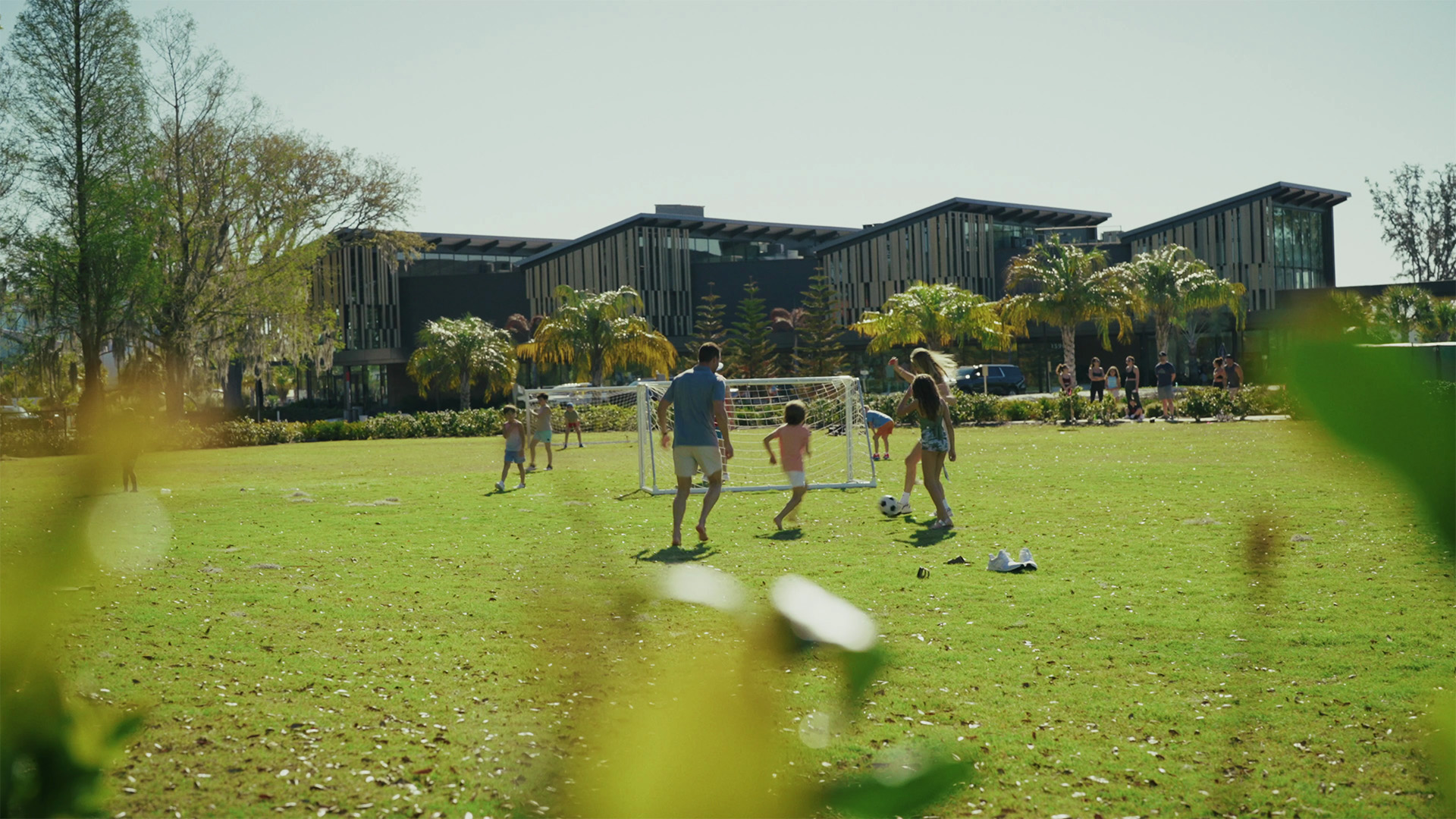 Group of people playing soccer on a sunny green field with modern buildings and palm trees in background.