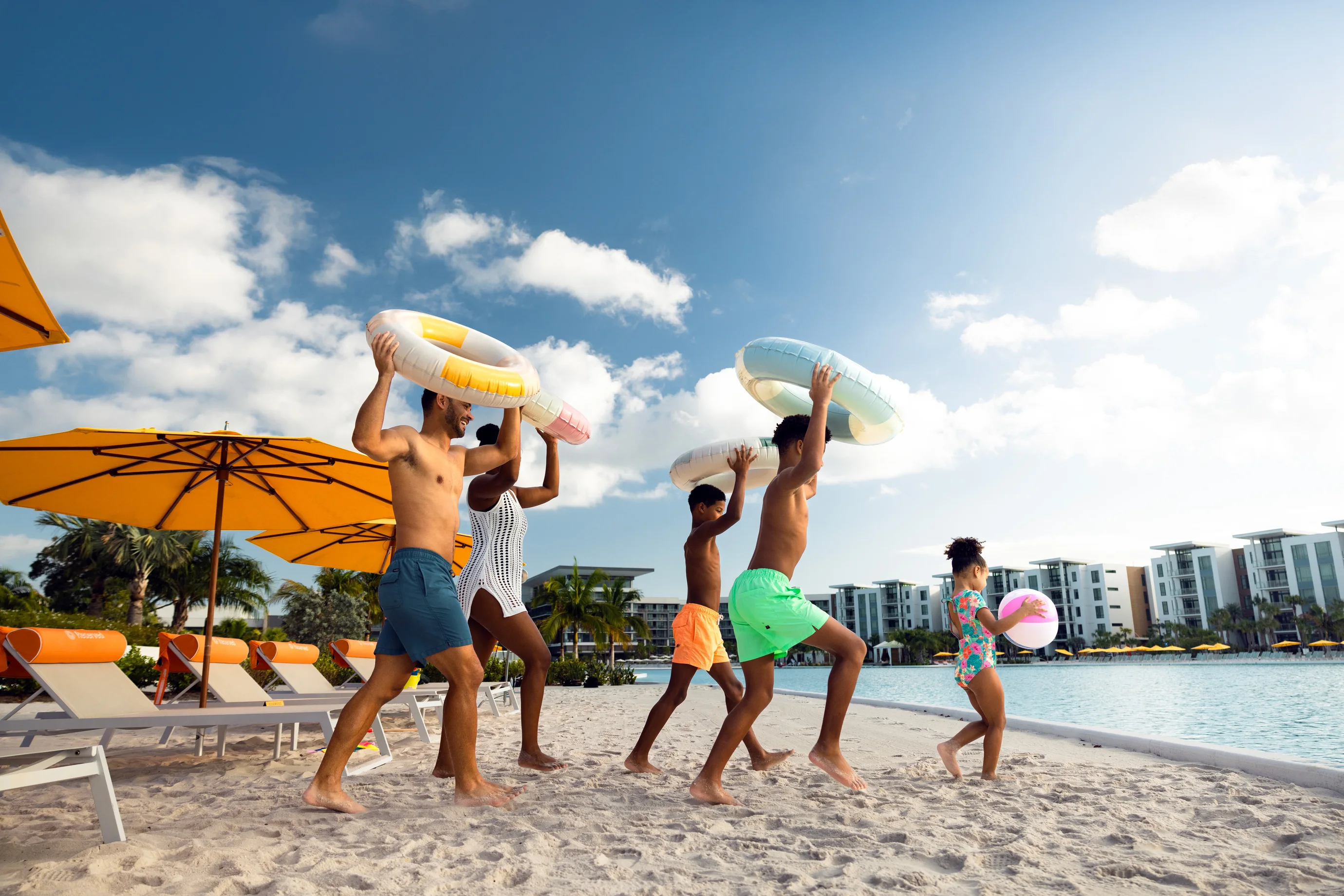Family walking on sandy poolside beach with inflatable rings and beach ball on a sunny day.