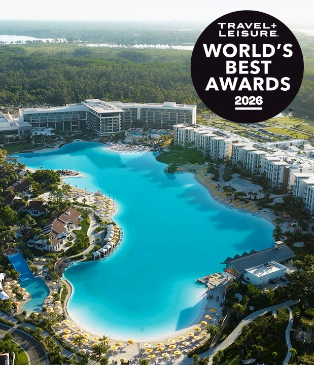 Aerial view of a large resort pool with buildings, surrounded by green trees and yellow umbrellas.