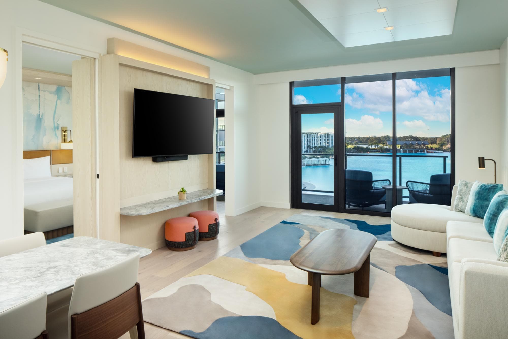 Bright suite with modern furniture, colorful rug, wall-mounted TV, and lake view through glass door.