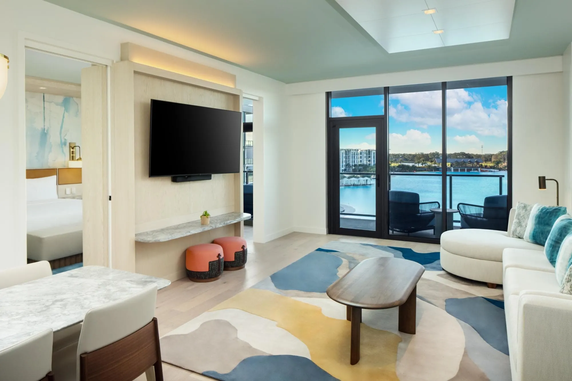 Bright suite with modern furniture, colorful rug, wall-mounted TV, and lake view through glass door.