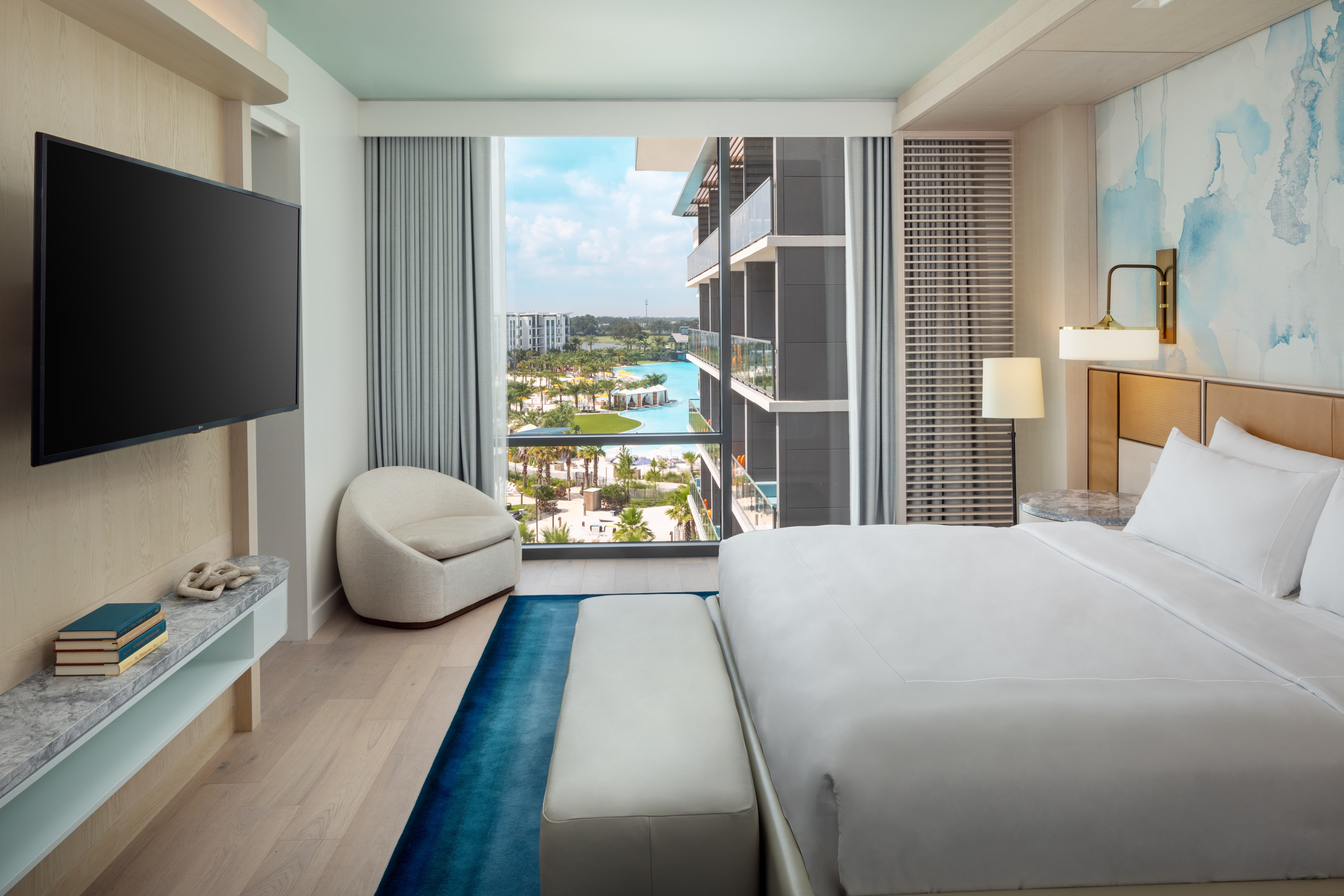 Modern hotel room with king bed, blue rug, round chair, TV, and large window overlooking pool and garden.