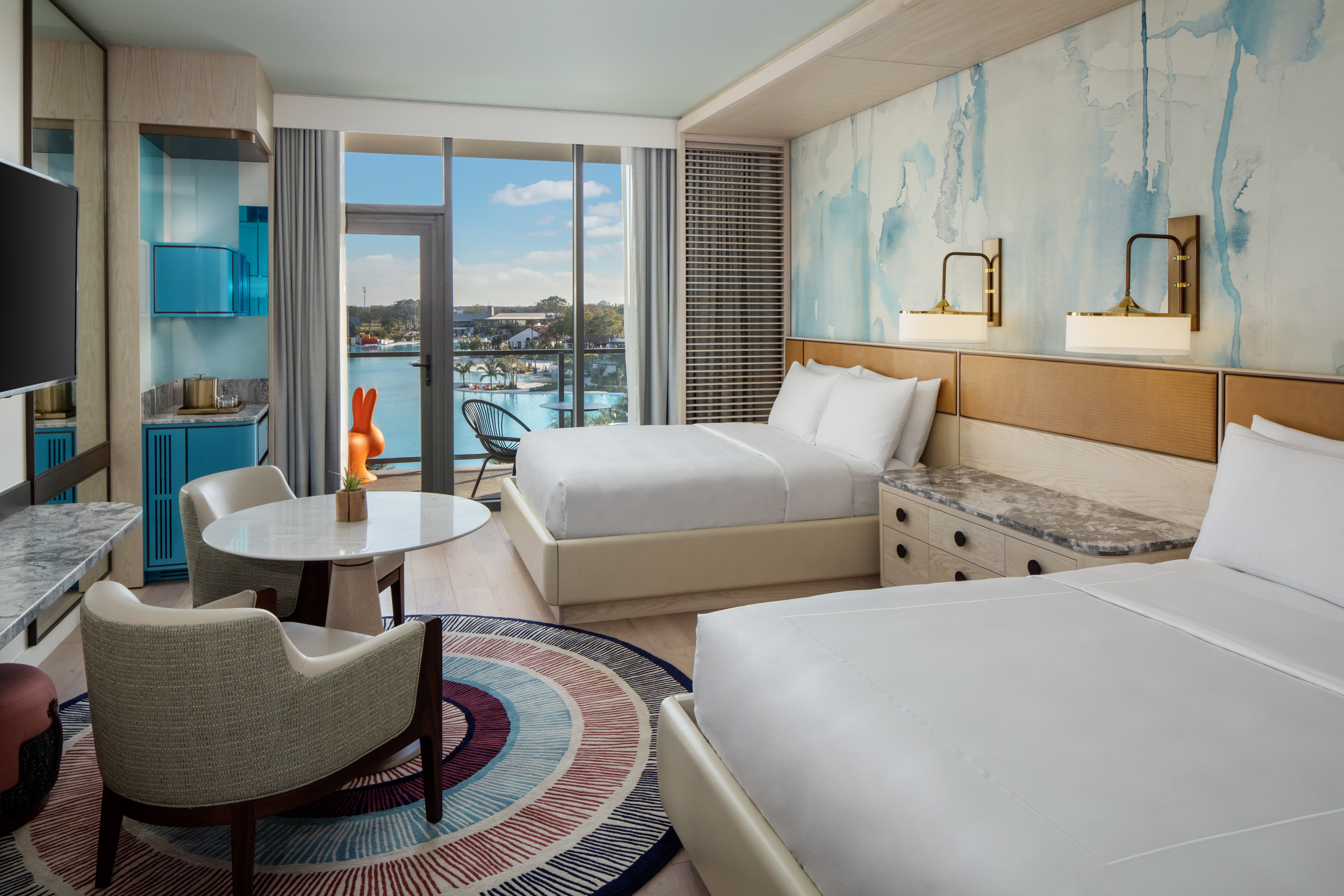 Hotel room with two beds, round table with chairs, balcony, and lagoon view outside glass doors.