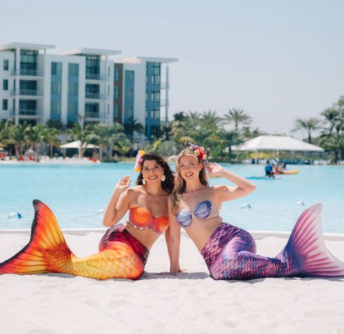 Two women with colorful mermaid tails sitting on white sand by a pool, waving and smiling.