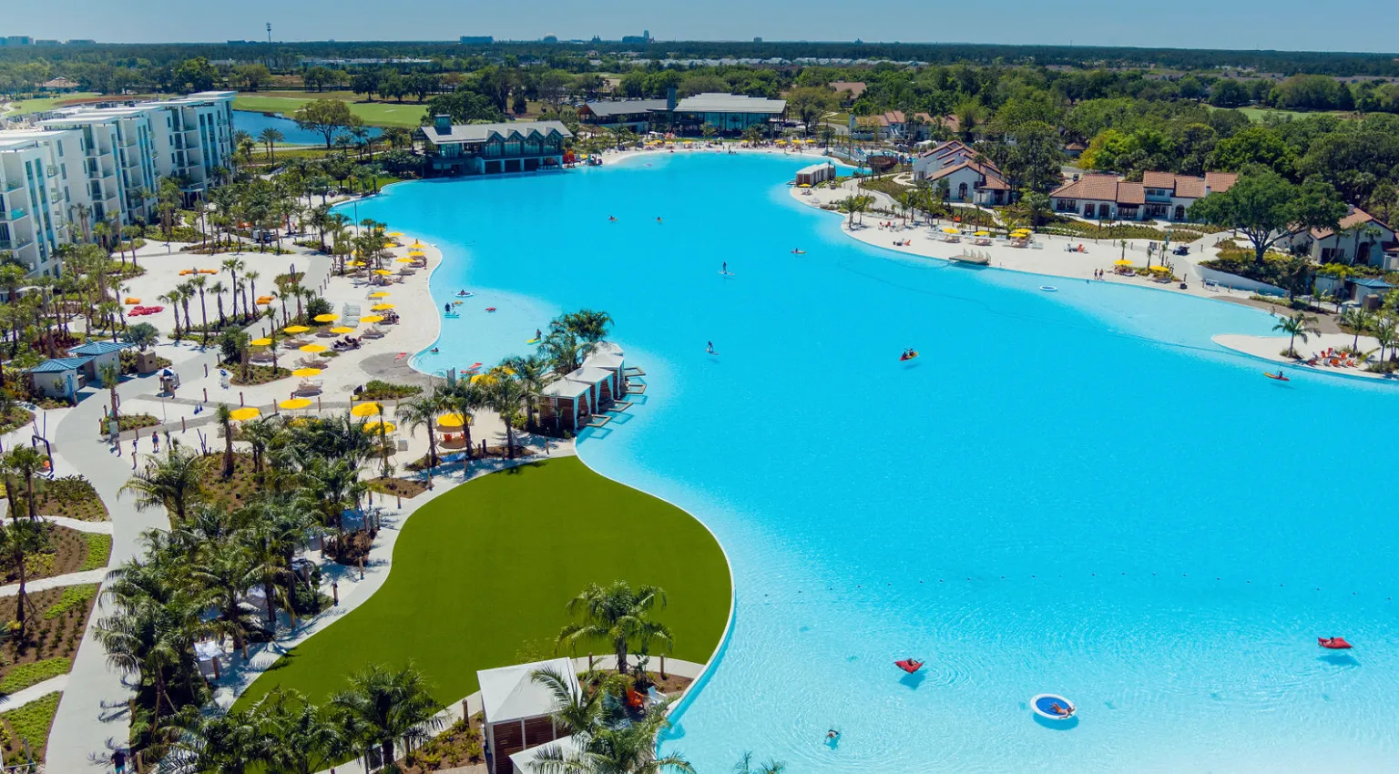 Large blue resort pool with people kayaking and lounging under yellow umbrellas by green landscaping.