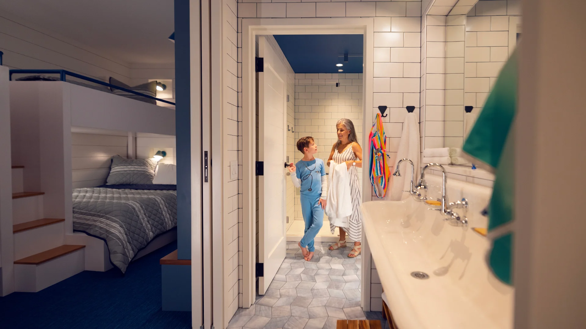 A child in blue pajamas talks with a woman holding towels in a modern bathroom near bunk beds.