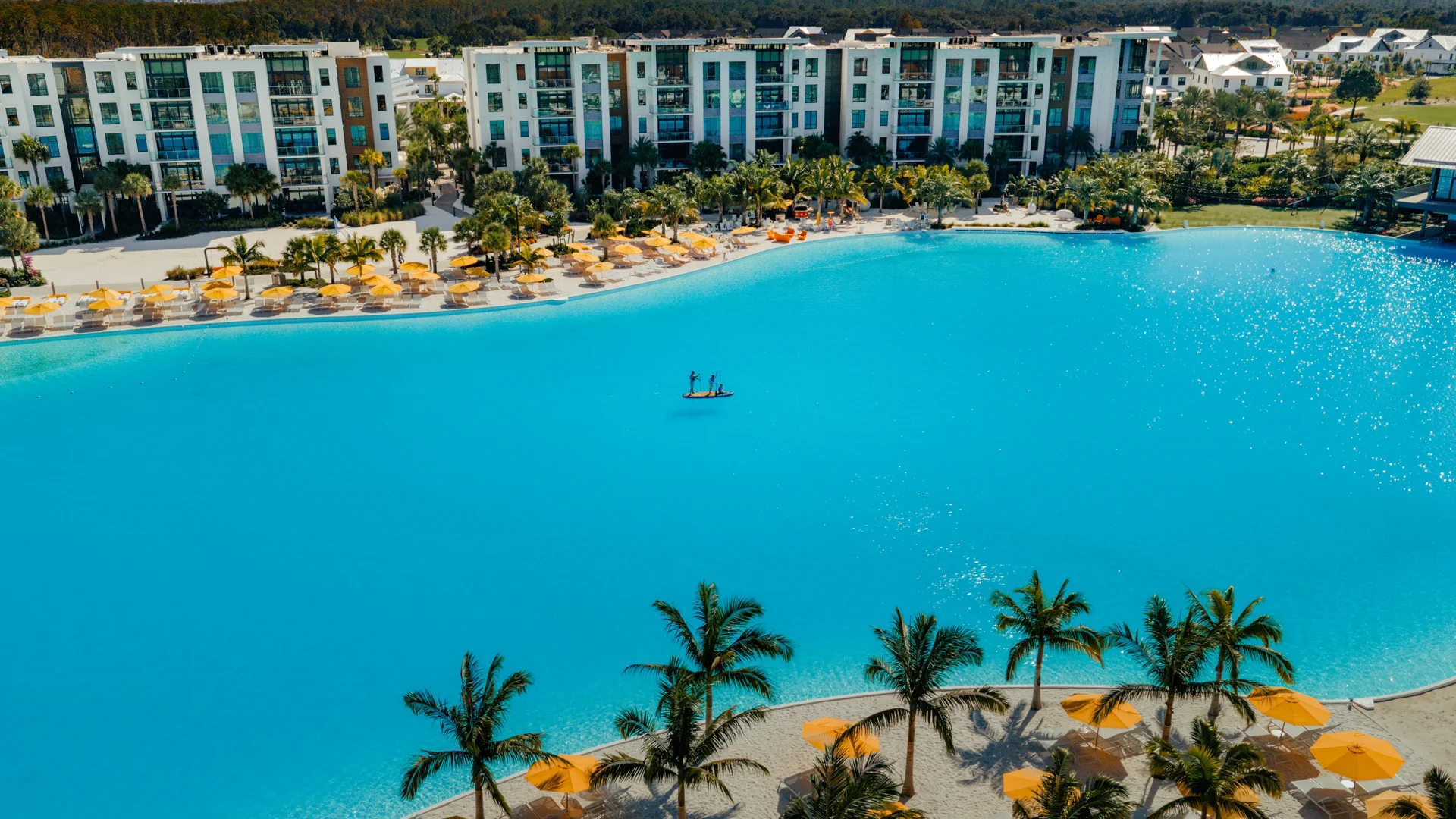 Large blue swimming lagoon with two paddleboarders, palm trees, yellow umbrellas, and modern white buildings.