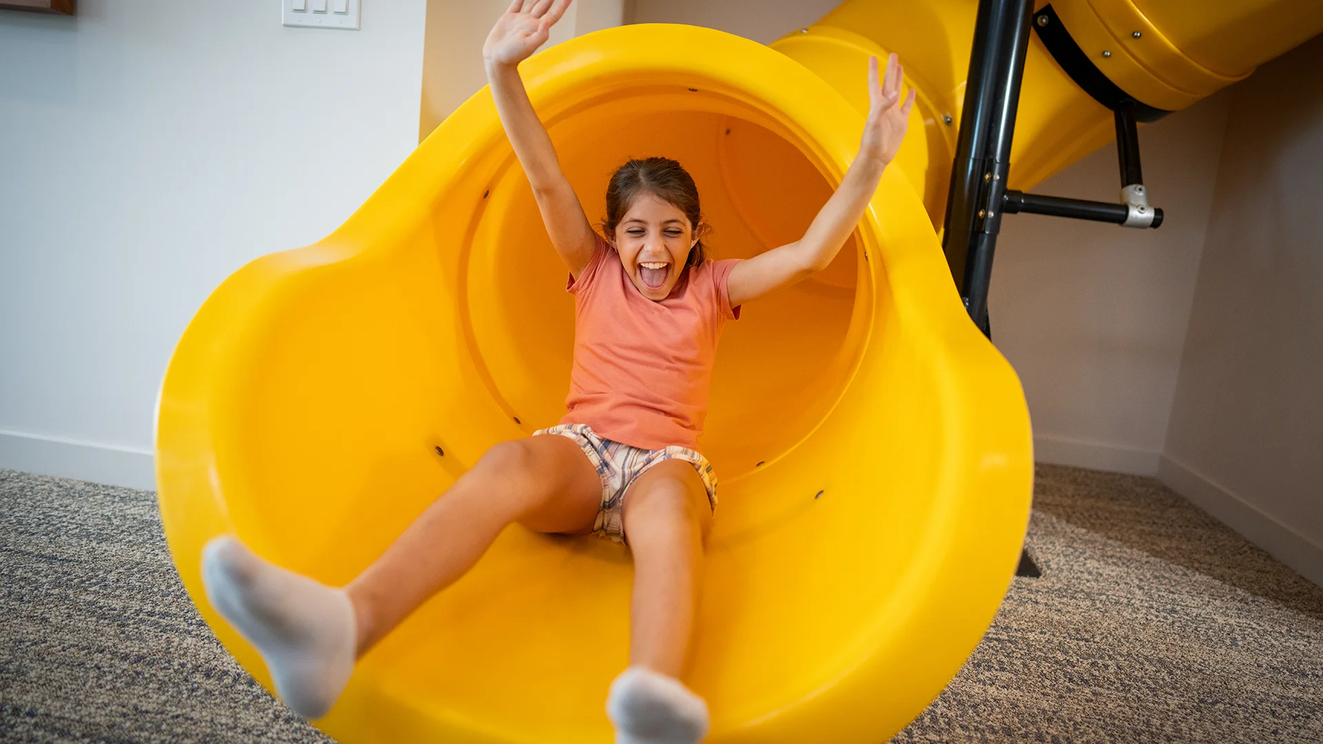 A happy young girl sliding down a bright yellow indoor slide with arms raised.