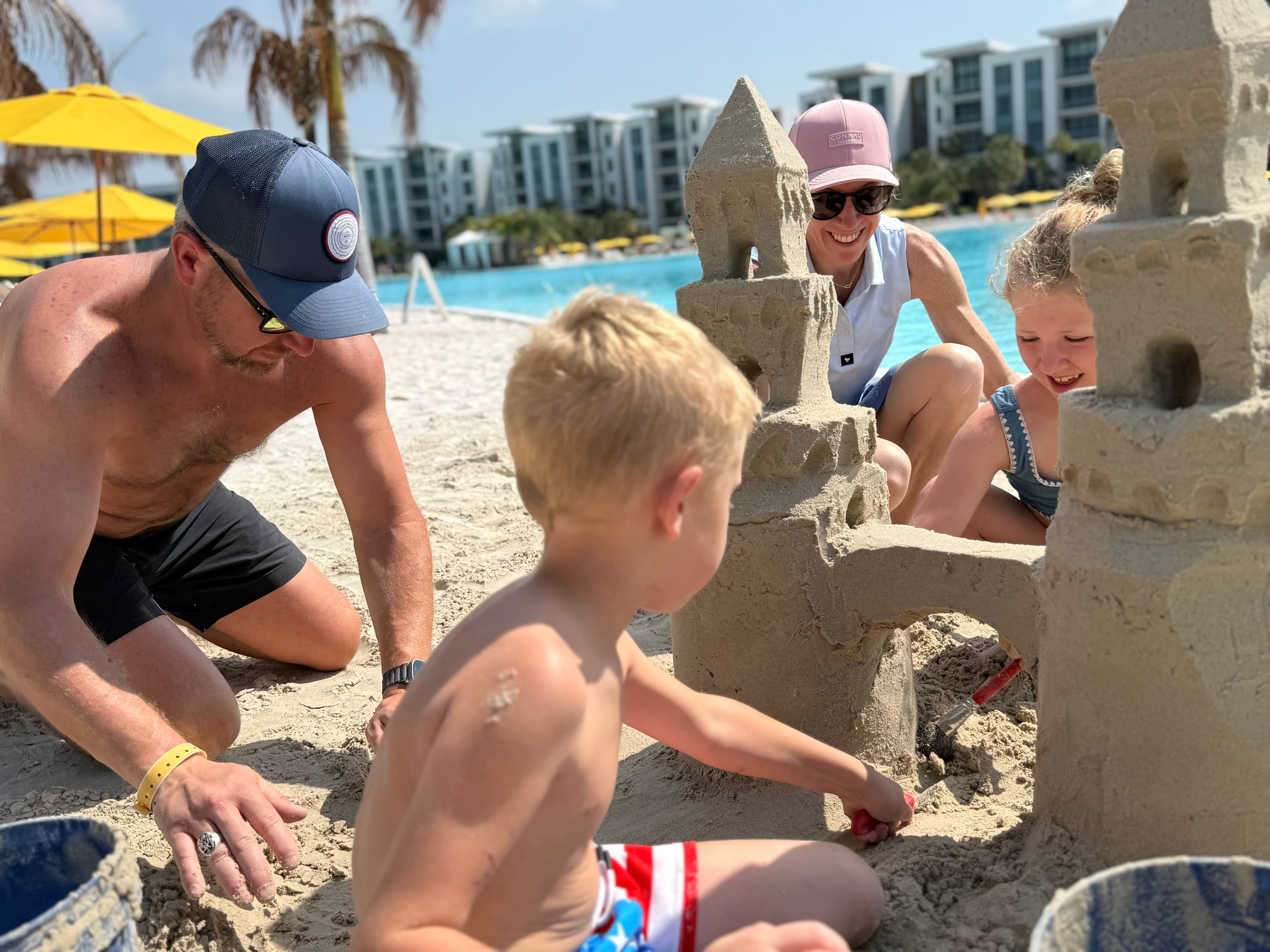 Family building a detailed sandcastle together on sandy beach beside a resort pool under blue sky.