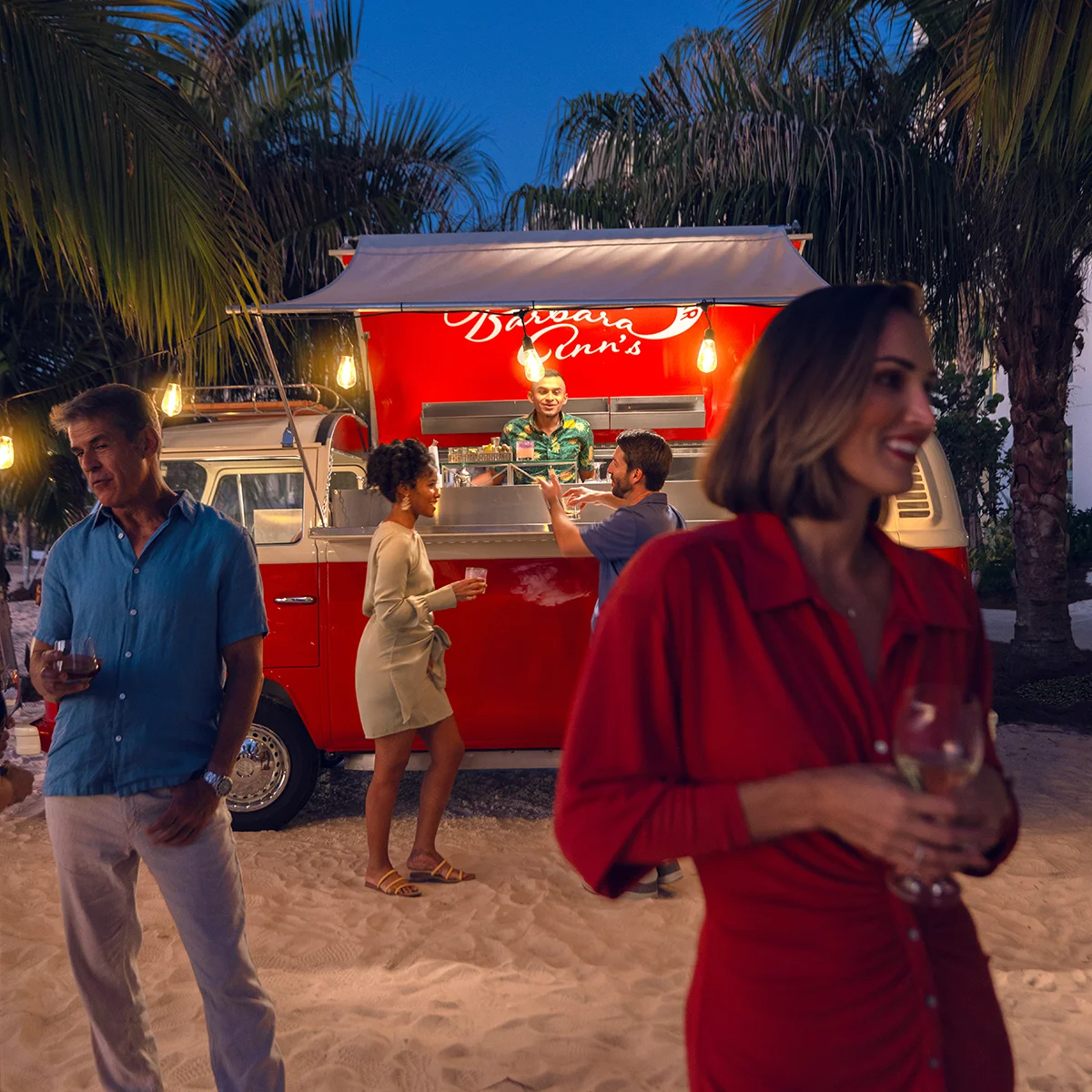 People enjoying drinks near a red food truck under palm trees at dusk on sandy ground.