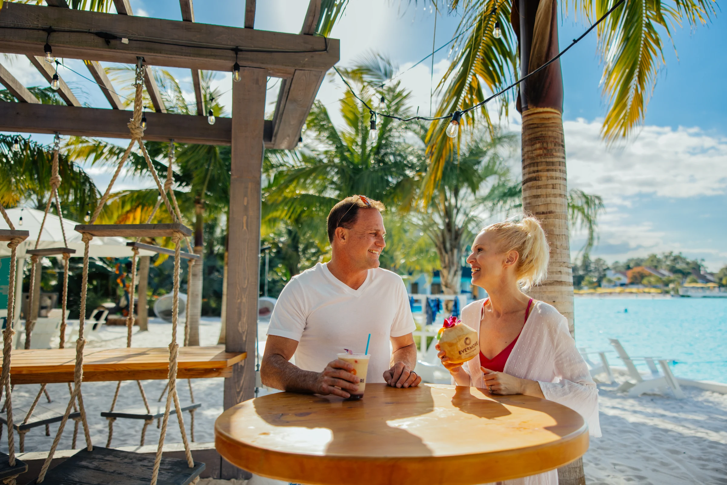 Smiling couple hold drinks at a wooden table on a sunny beach with palm trees and water view.