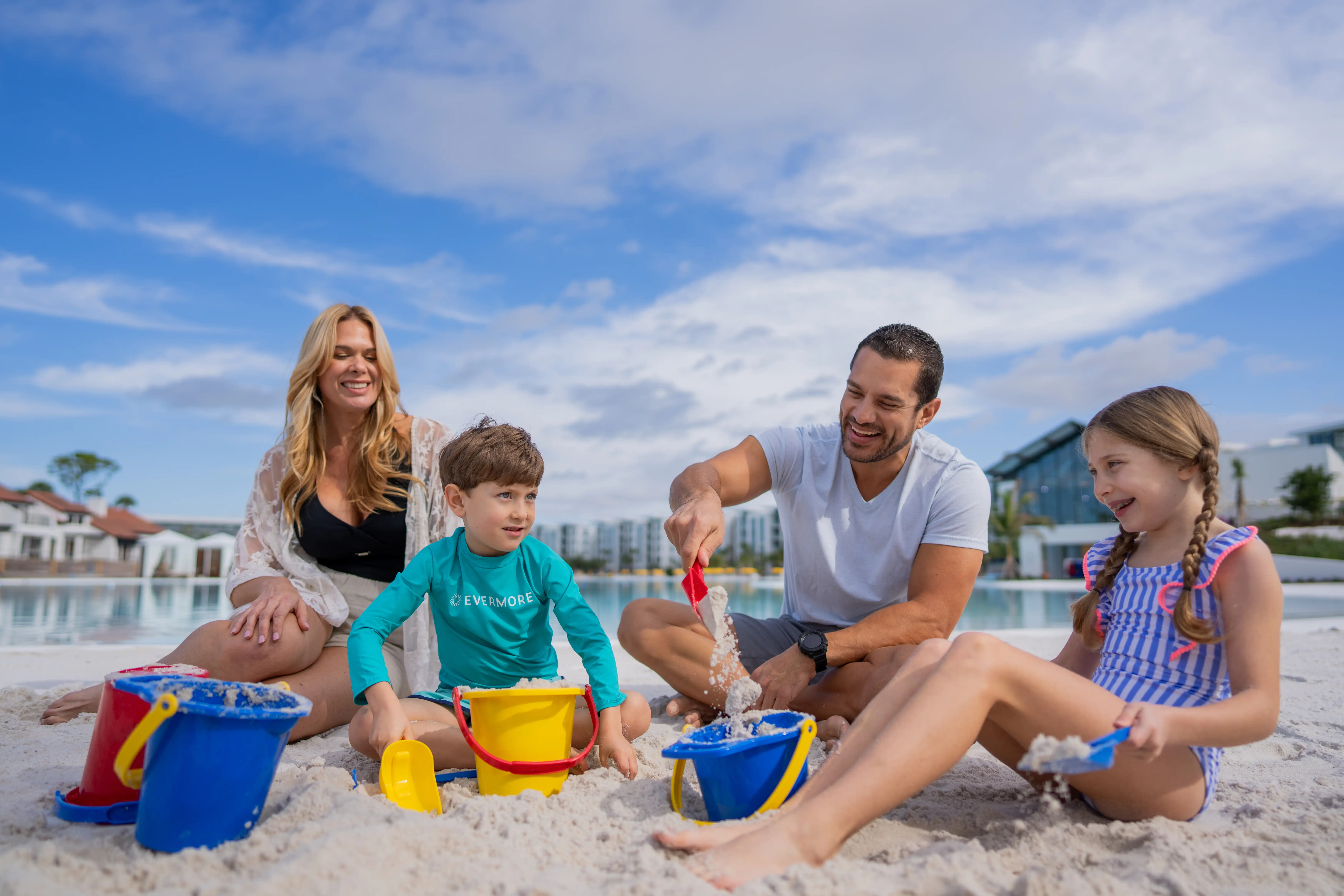 Family of four playing with buckets and shovels on a sandy beach near a pool under a blue sky.