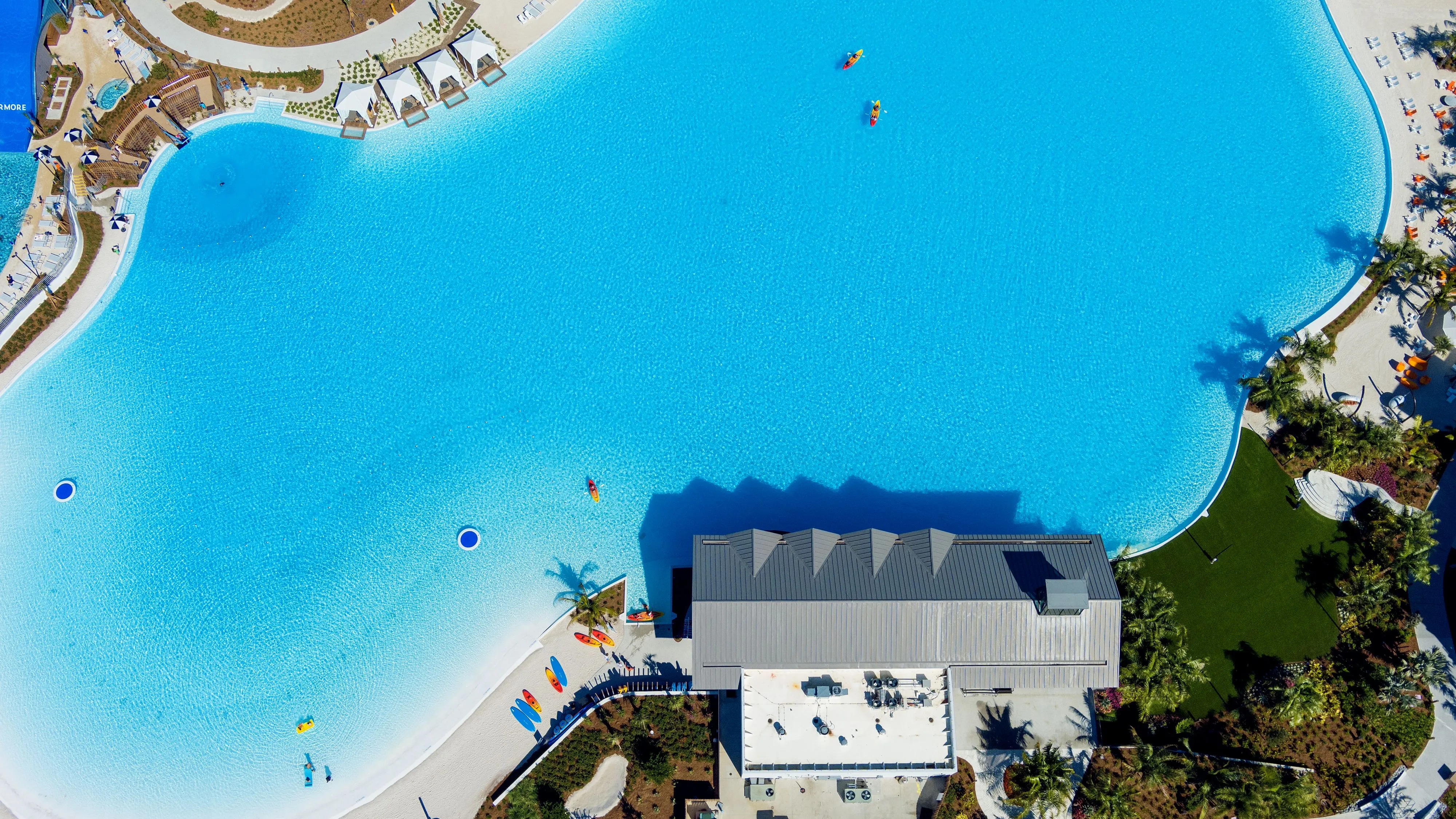 Aerial view of a large bright blue swimming pool with kayaks on the water and sandy beach areas with lounge chairs around it.