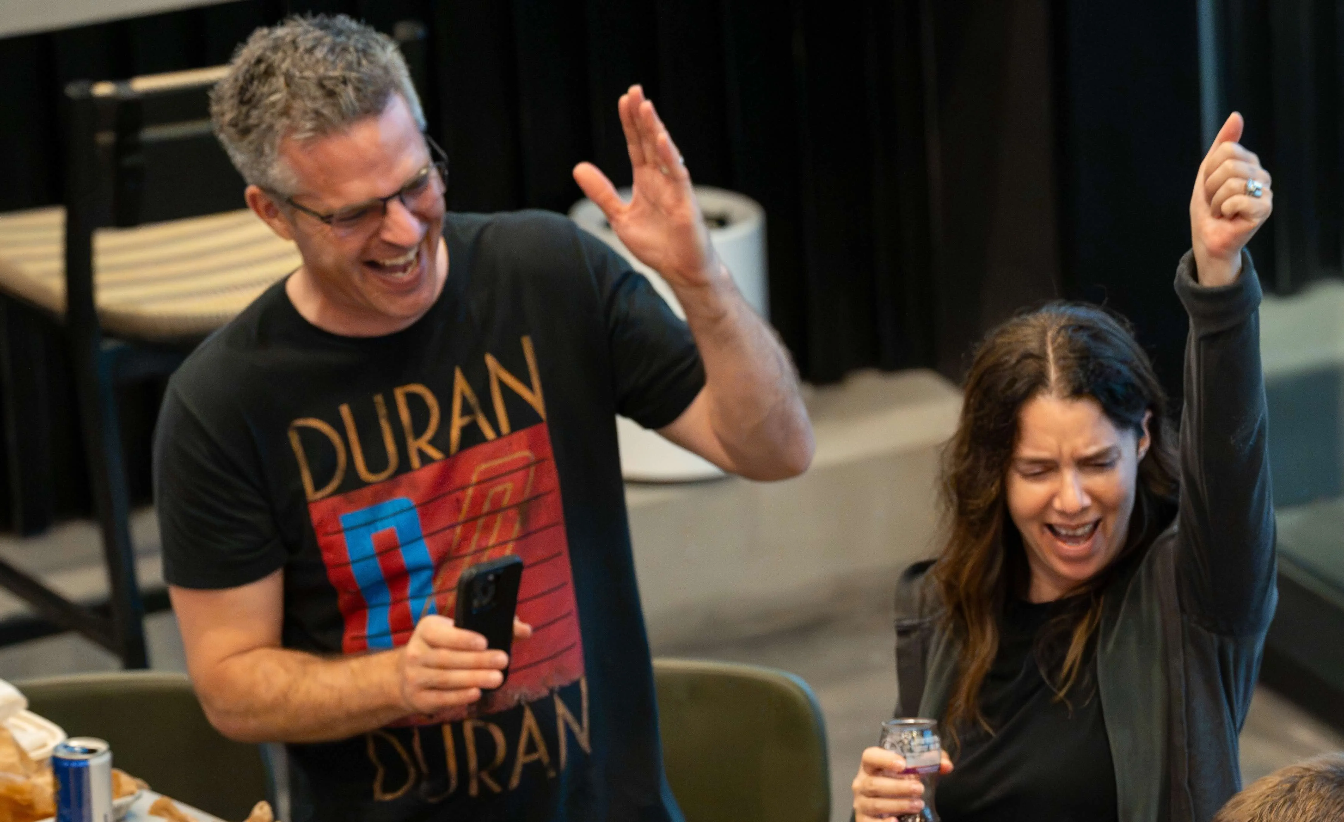 A man wearing glasses and a Duran Duran t-shirt and a woman with long dark hair raise their hands in celebration indoors, the woman holding a can and the man holding a phone, both smiling and cheering.