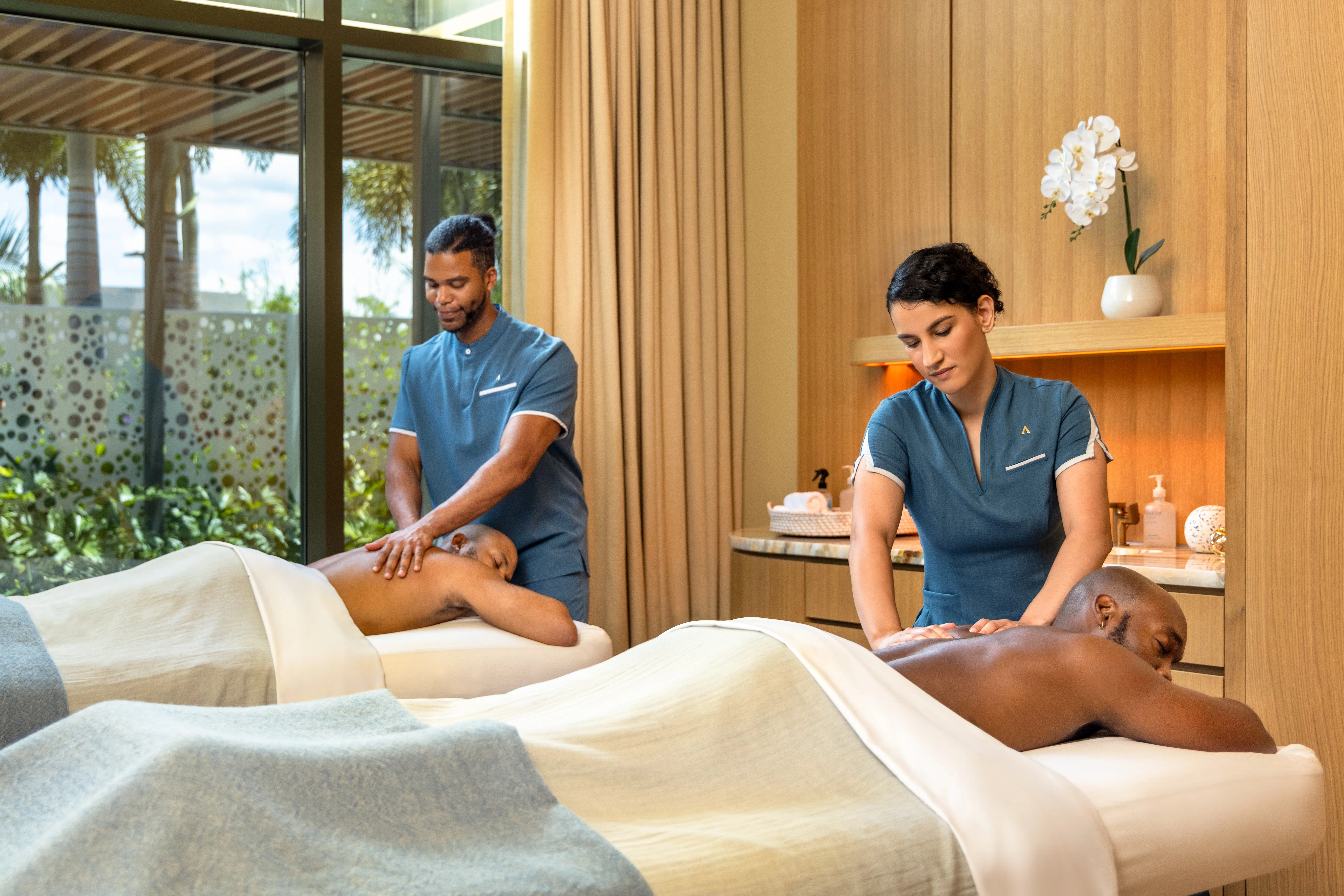 Two massage therapists in blue uniforms giving back massages to two men lying on massage tables covered with light-colored sheets in a spa room with wooden walls and a window showing greenery outside.