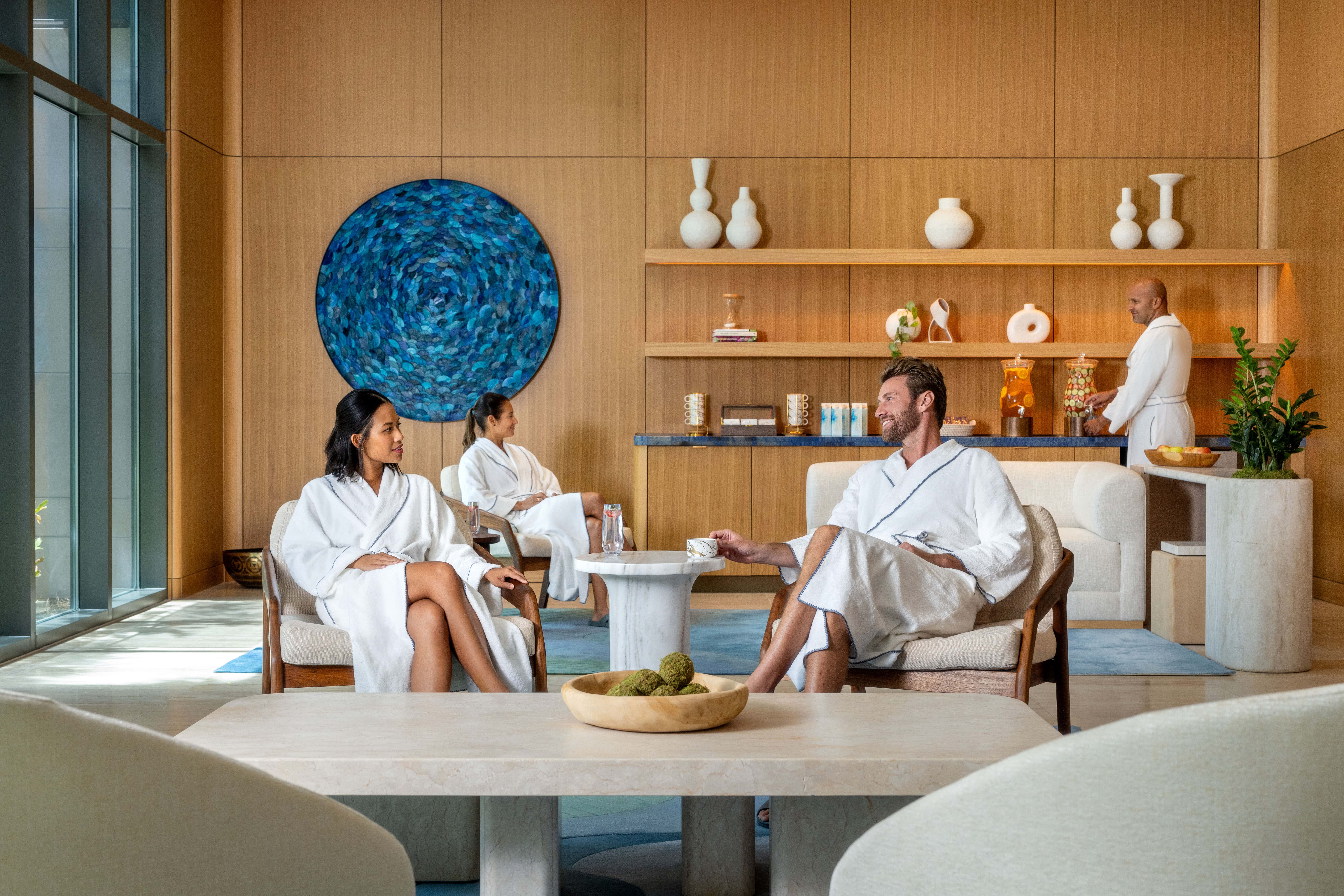 A modern spa lounge with four people wearing white robes; two women seated on chairs on the left, one man seated on a chair on the right holding a teacup, and a man standing near a refreshments counter with infused water dispensers. The background features wood panel walls, white decorative vases on shelves, a large blue circular wall art, and green plants.