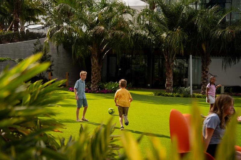 Children playing soccer on a lawn surrounded by tropical landscaping.