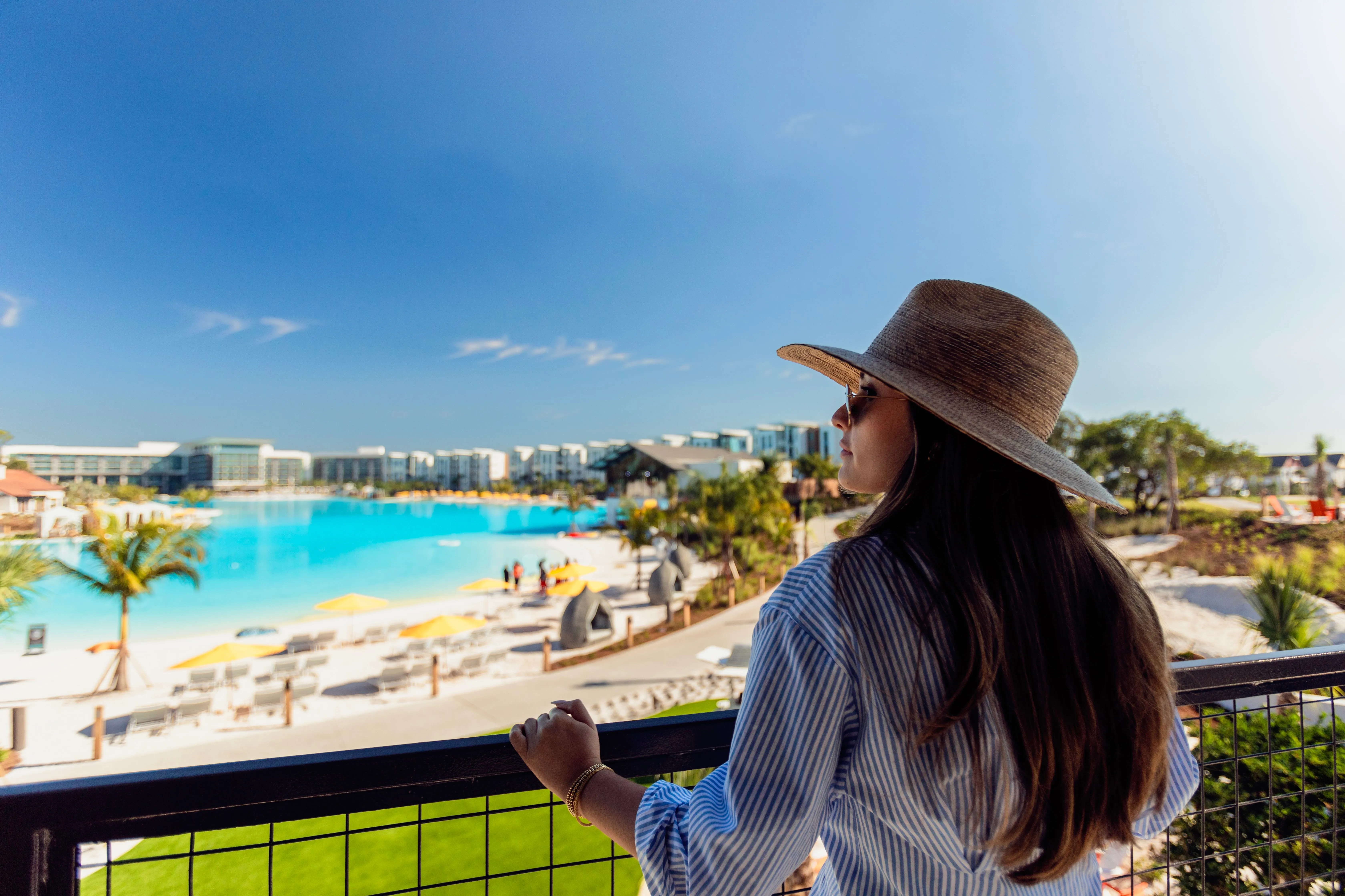 Woman wearing a wide-brimmed hat and sunglasses looks out over a large pool with yellow umbrellas and lounge chairs on a sunny day at a resort.