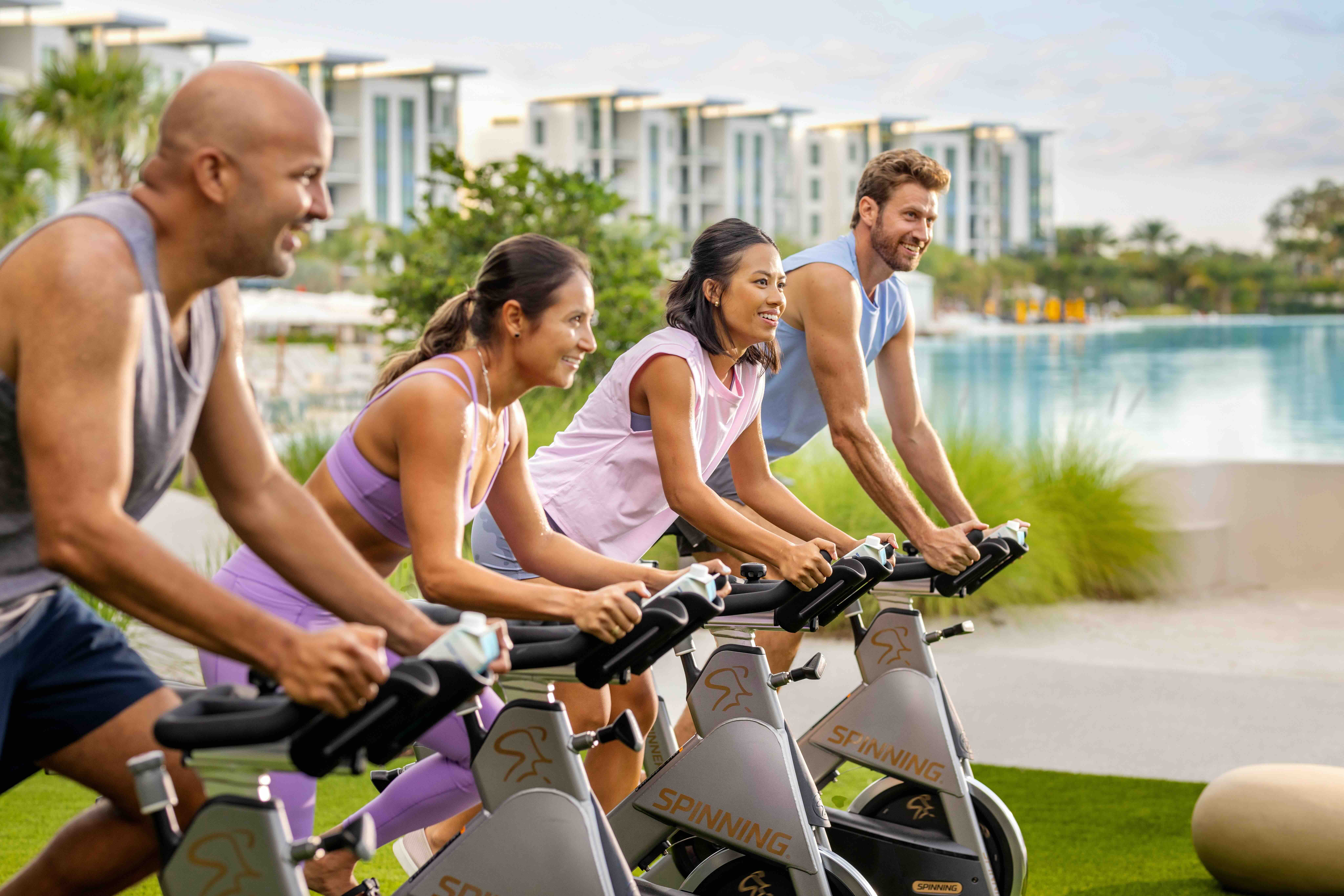 Four diverse adults smiling and cycling on stationary spin bikes outdoors on green grass near a modern building and a body of water during daylight.