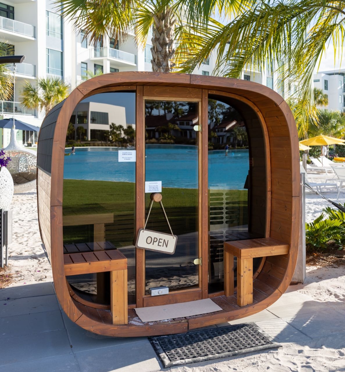 Outdoor wooden sauna with dark glass doors and 'OPEN' sign hanging on door handle, set near a poolside with palm trees, white sandy area, lounge chairs, umbrellas, and modern white buildings in the background.