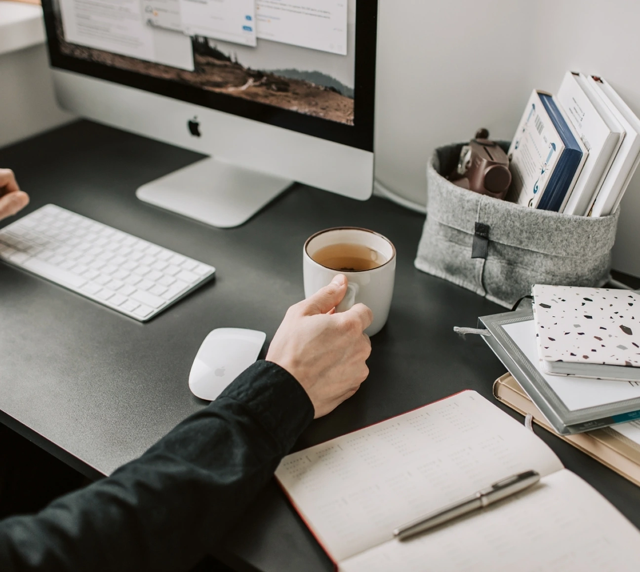 Person's hand holding a coffee mug beside an Apple keyboard, mouse, notebooks, and computer on desk.