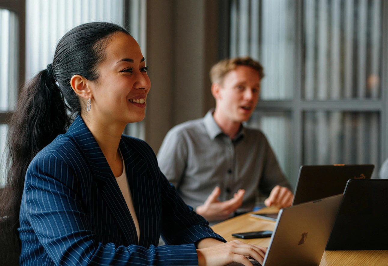 Smiling woman in pinstripe blazer using a laptop during a discussion with a man in the background.