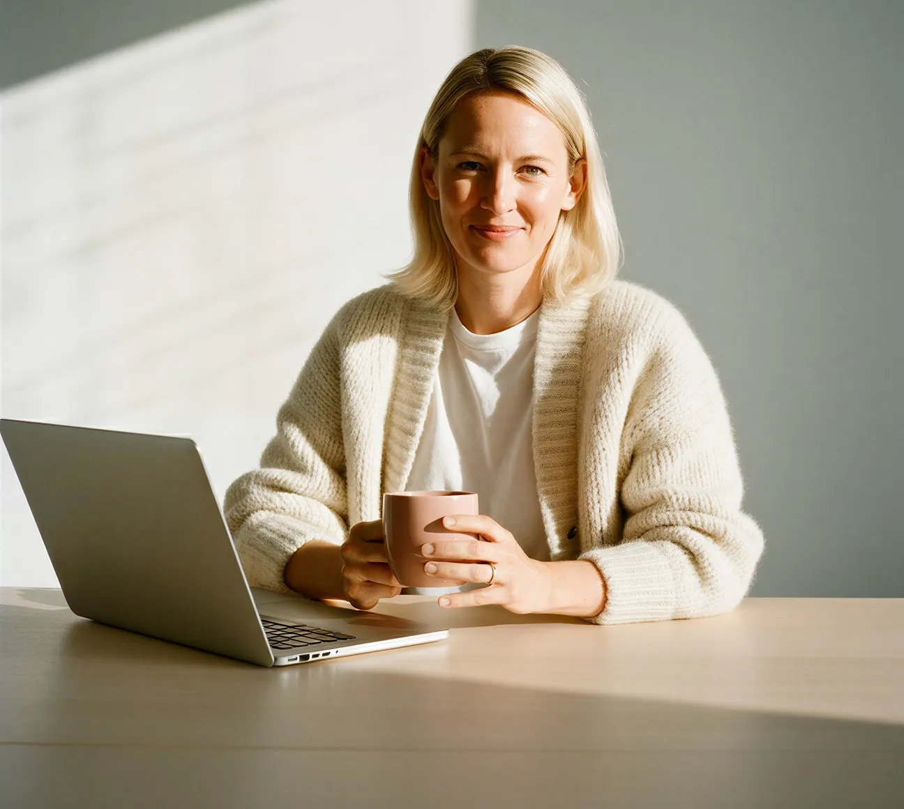 Smiling woman in cream sweater holding a mug sitting at a table with a laptop in soft daylight.