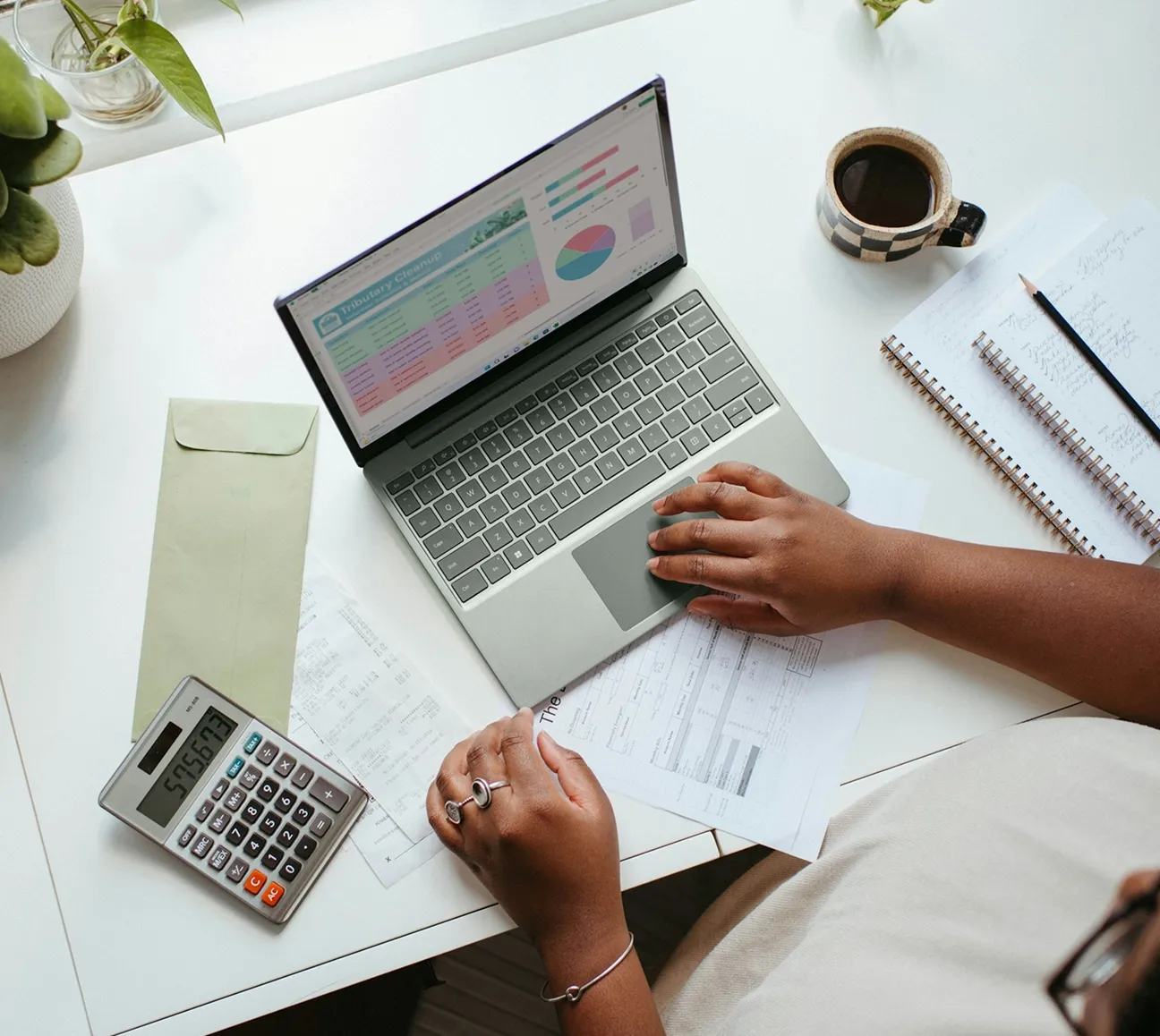 Person using laptop on a white desk with calculator, papers, notebooks, pencil, coffee, and plants.