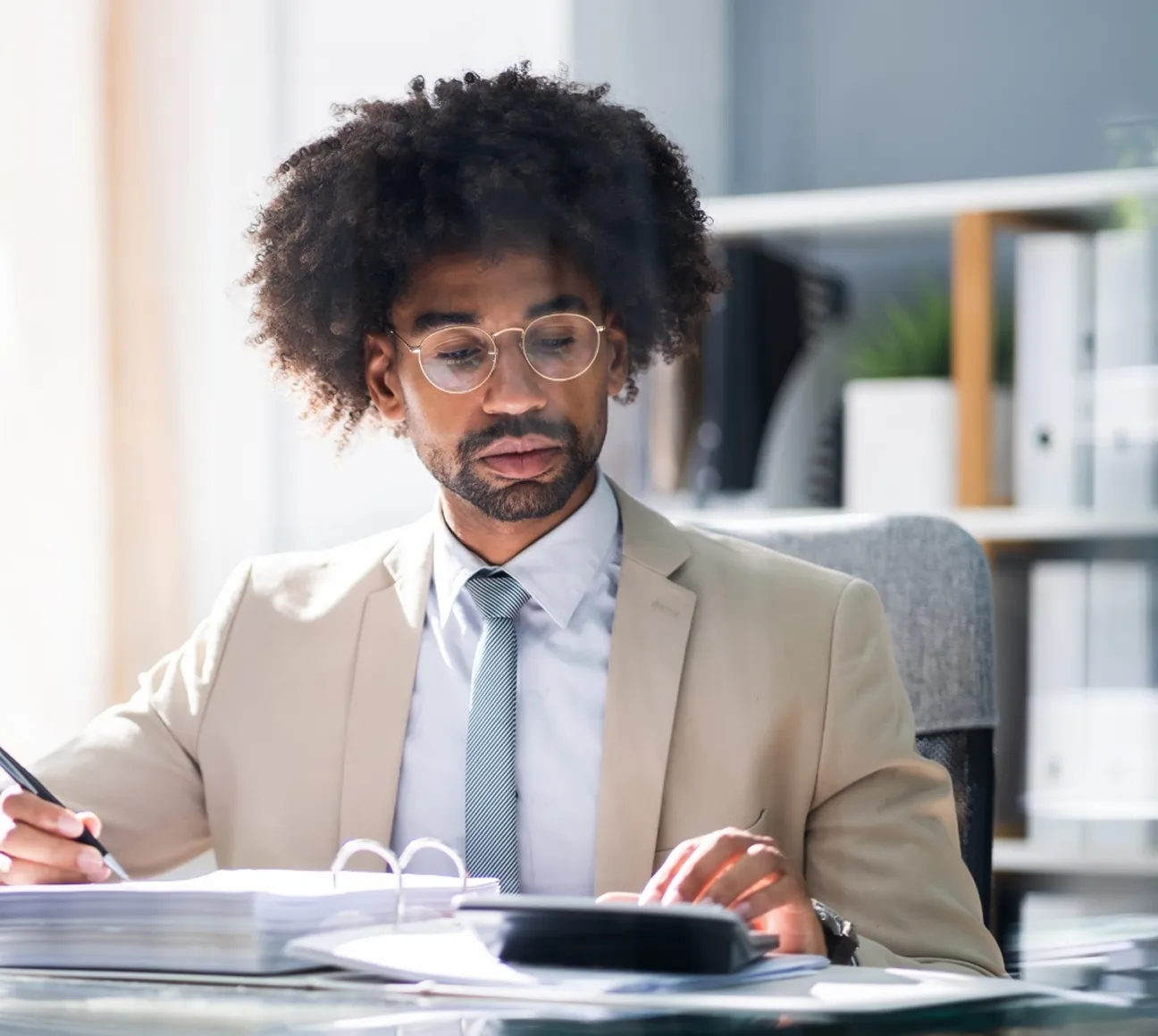 Man with curly hair and glasses writing in a notebook at a desk in a bright office.