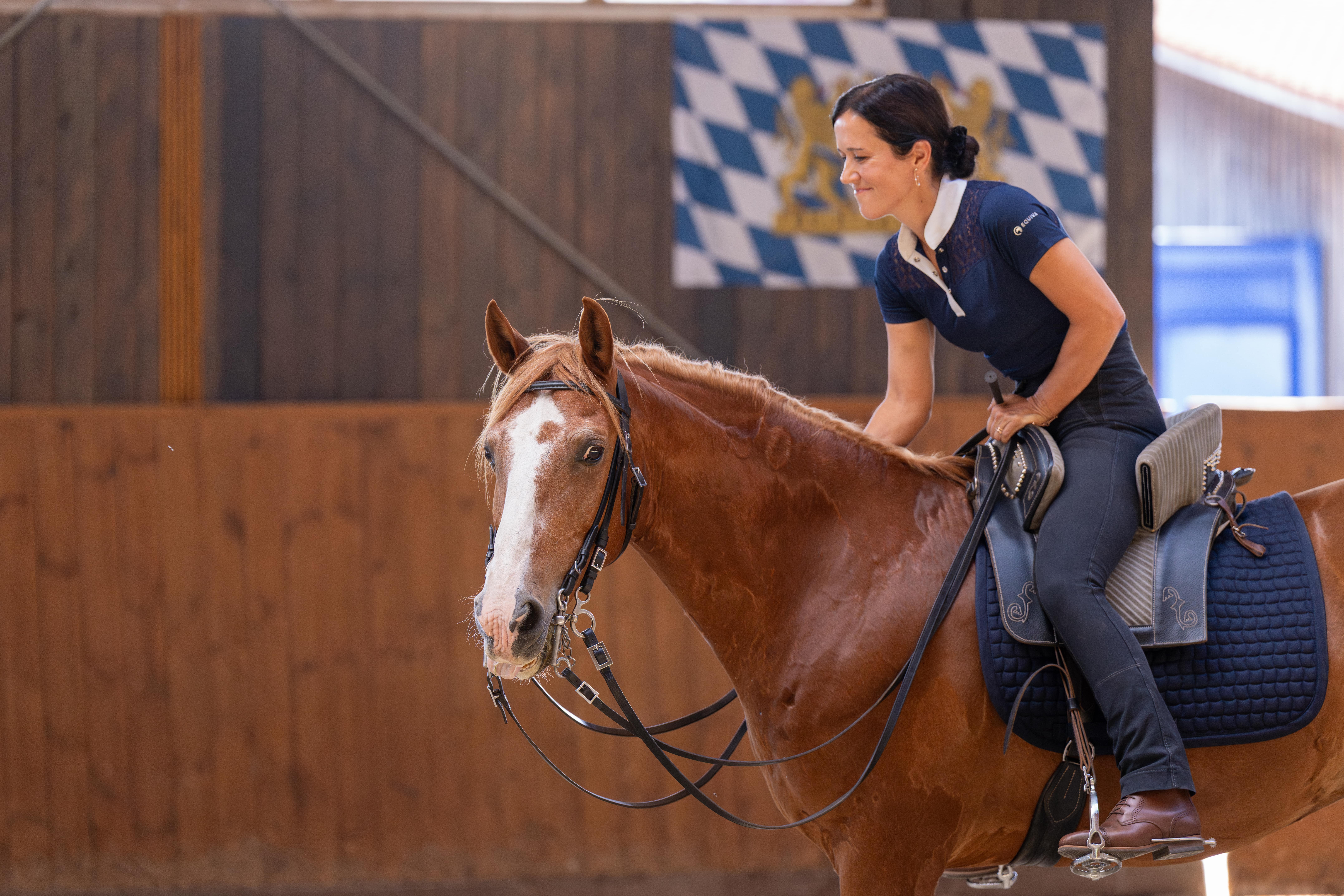 Frau reitet ein braunes Pferd in einer Reithalle mit bayerischer Flagge im Hintergrund.