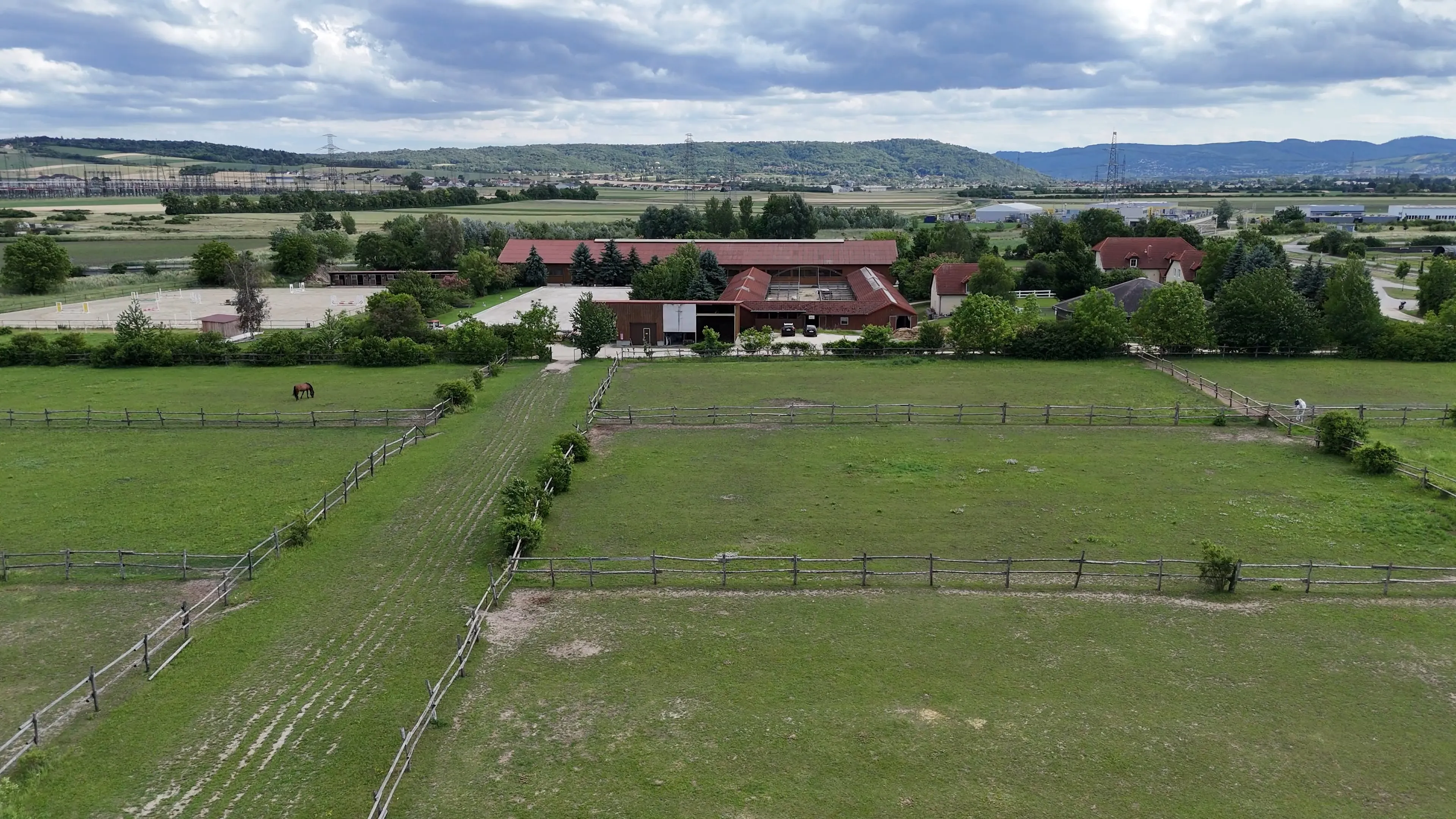 Ländliche Pferdefarm mit umzäunten grünen Weiden, Gebäuden und bewölktem Himmel im Hintergrund.