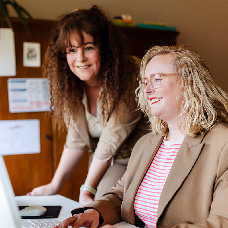 Carolien en Jolien kijken lachend samen naar een computerscherm in hun praktijk