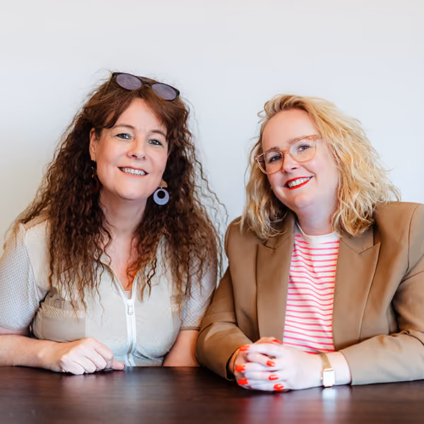 Twee vrouwen zitten aan een tafel, glimlachen naar de camera met casual kleding en bril.