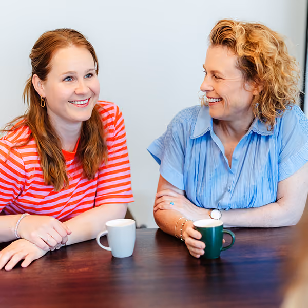 Twee vrouwen zitten tegenover elkaar en praten terwijl ze koffie drinken aan een houten tafel.