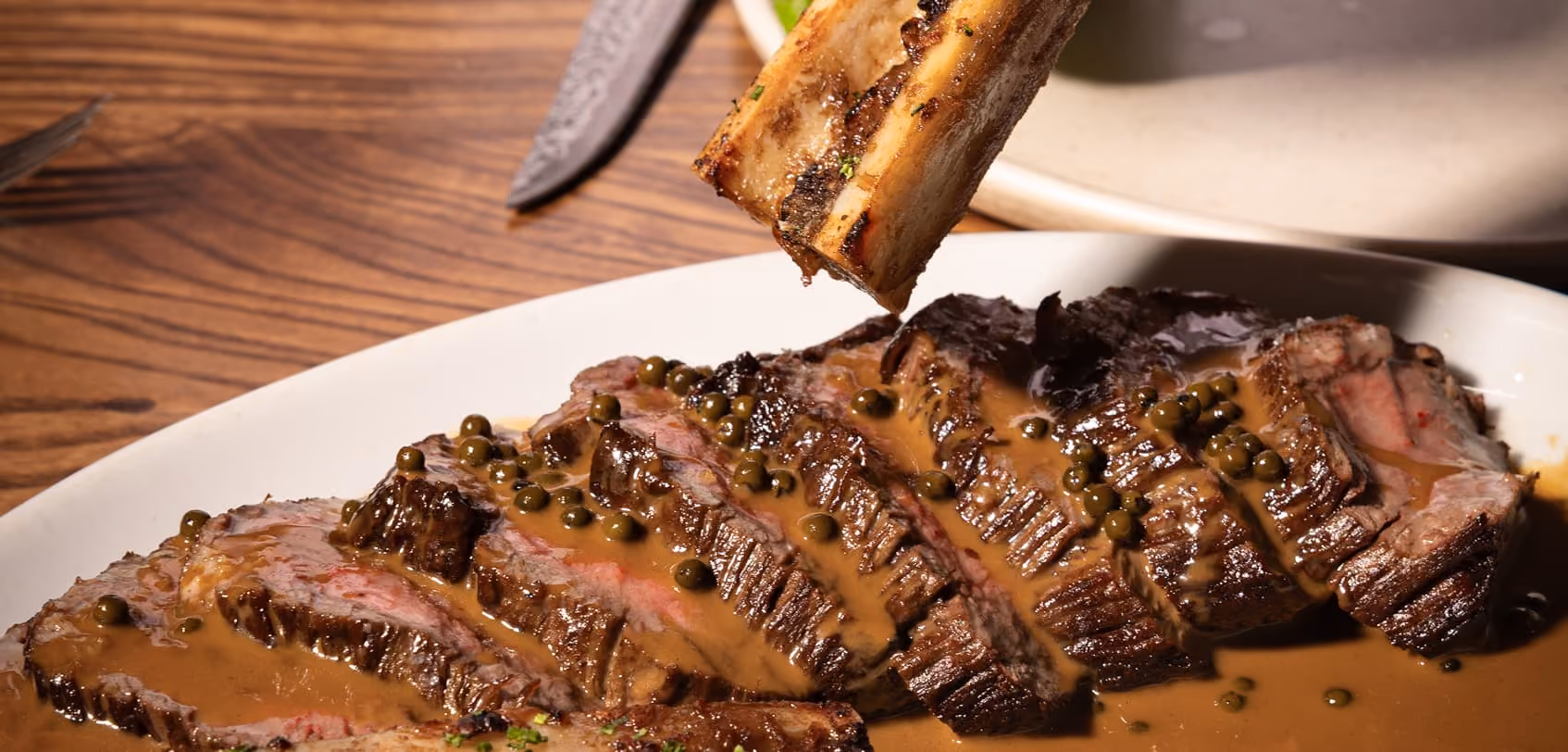 Sliced steak on plate topped with brown sauce and green peppercorns with a marrow bone above.