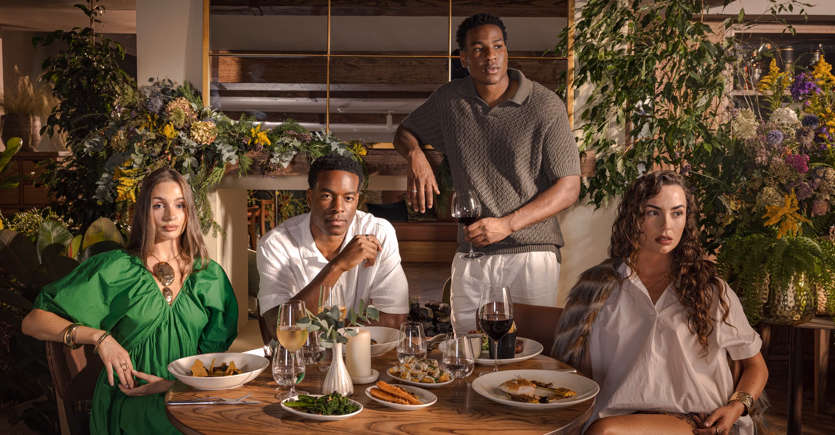 Two men and two women at a table with plates of food and wine glasses in a green floral setting.