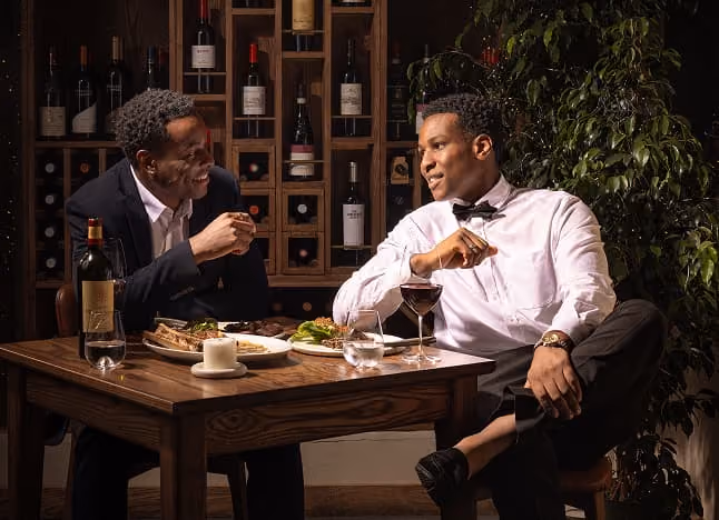 Two men dressed formally enjoy a meal and wine while conversing at a wooden table in a cozy setting.