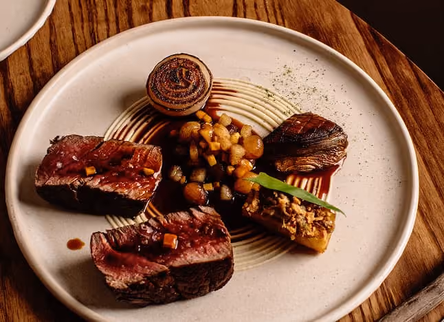 A gourmet plate with two slices of seared steak, grilled onion, sauce, diced vegetables, and a garnish on cream puree.