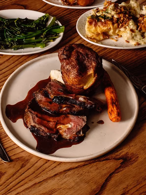 Plate with sliced roast beef, Yorkshire pudding, carrot, and two side dishes on wooden table.